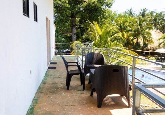 Balcón con sillas y mesa negras, pared blanca y exuberantes árboles tropicales.