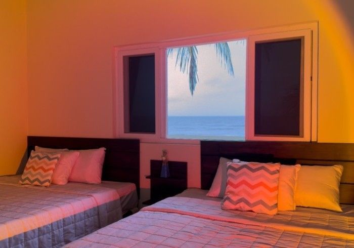 Dos camas en una habitación con vista al mar, iluminada por un atardecer naranja.