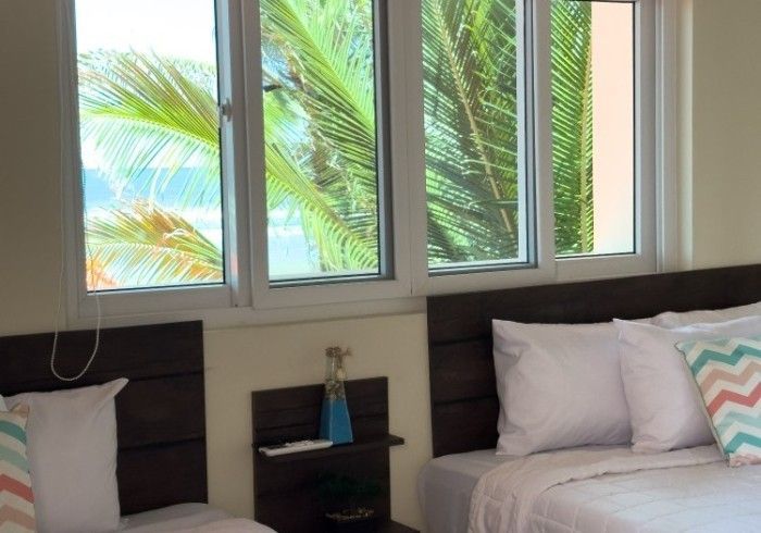 Dormitorio con ropa de cama blanca, cabeceros de madera oscura y una ventana con vista a las palmeras.