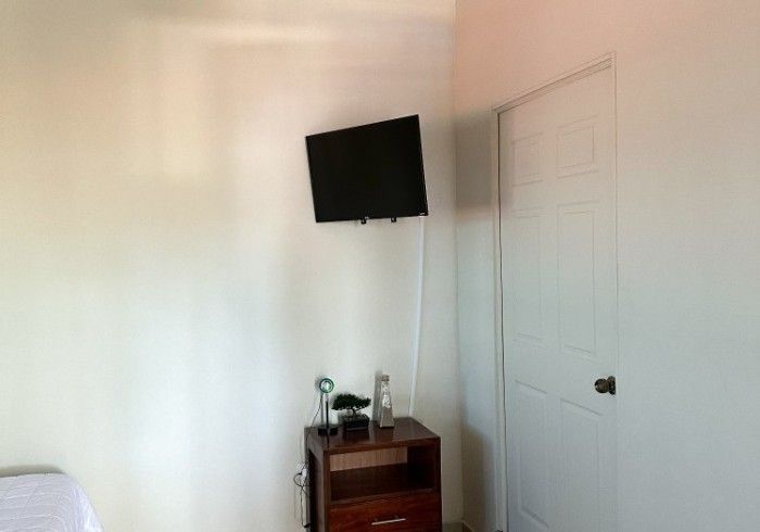 Dormitorio con televisor montado sobre una mesita de noche y una puerta blanca cerrada.