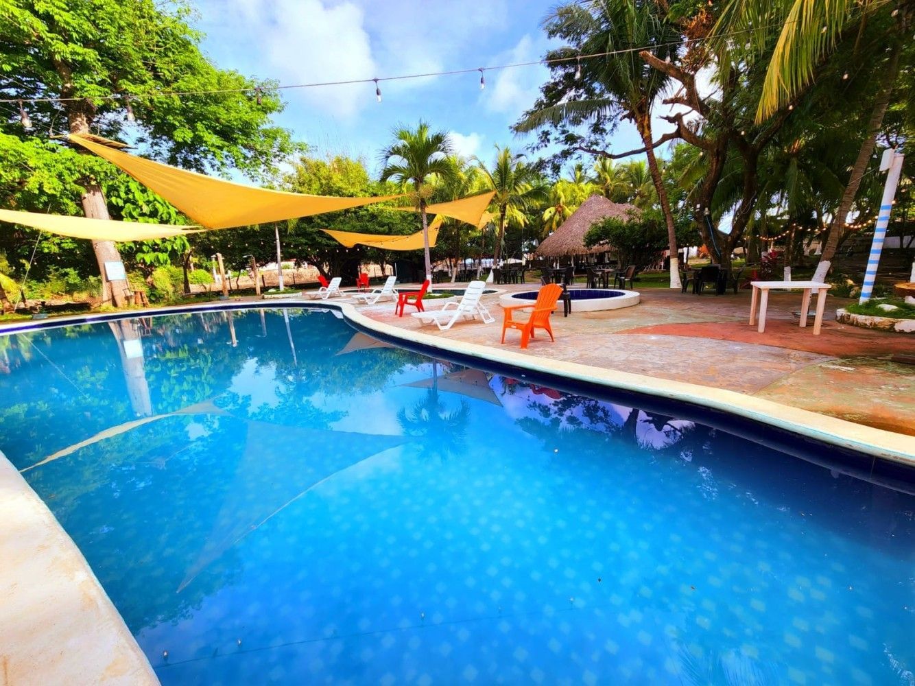 Una piscina azul con tumbonas, una estructura con techo de paja y árboles bajo un cielo soleado.