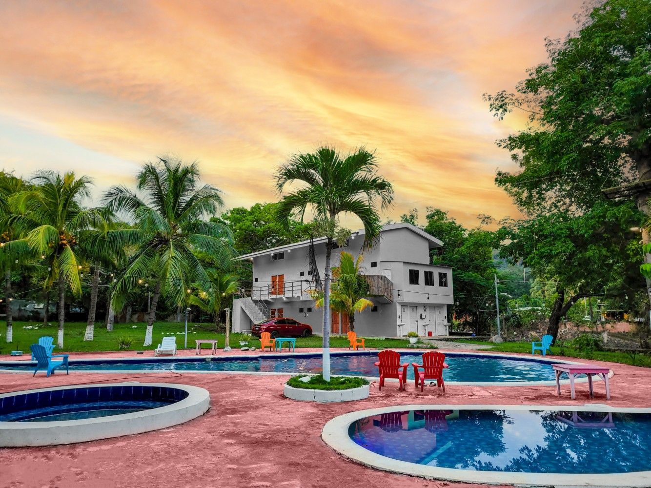 Un resort con zona de piscina, palmeras y un edificio bajo un cielo anaranjado al atardecer.
