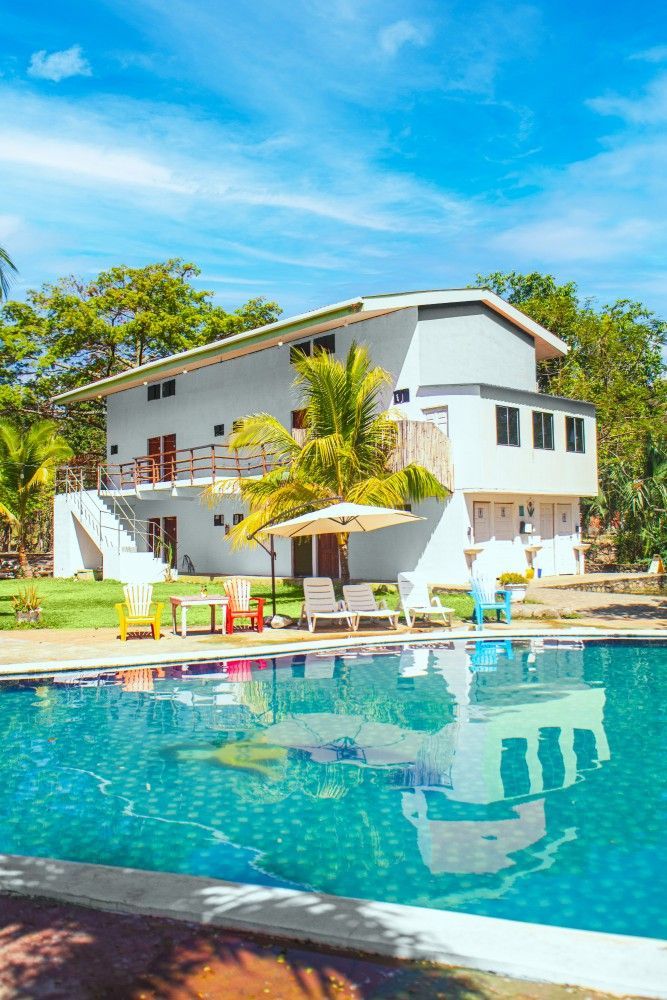 Edificio blanco de dos pisos con piscina al frente, sillas y cielo soleado.