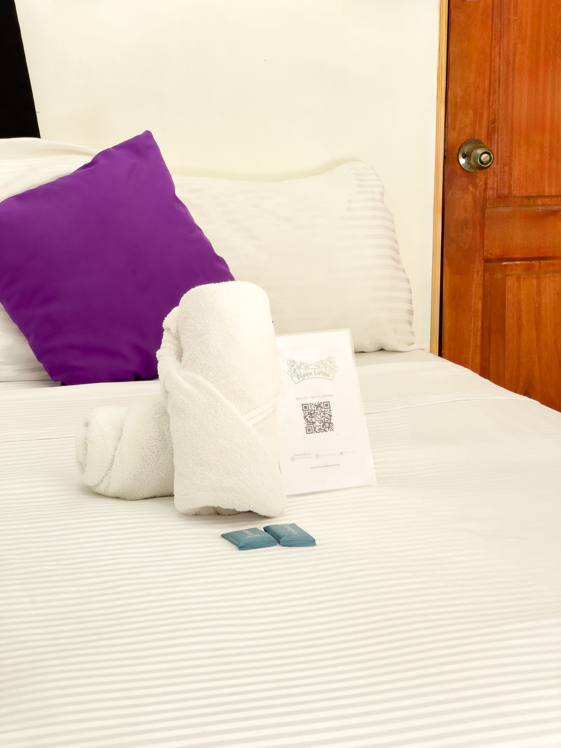 Cama con sábanas blancas, toallas enrolladas, almohada morada y hoja de información de la habitación.