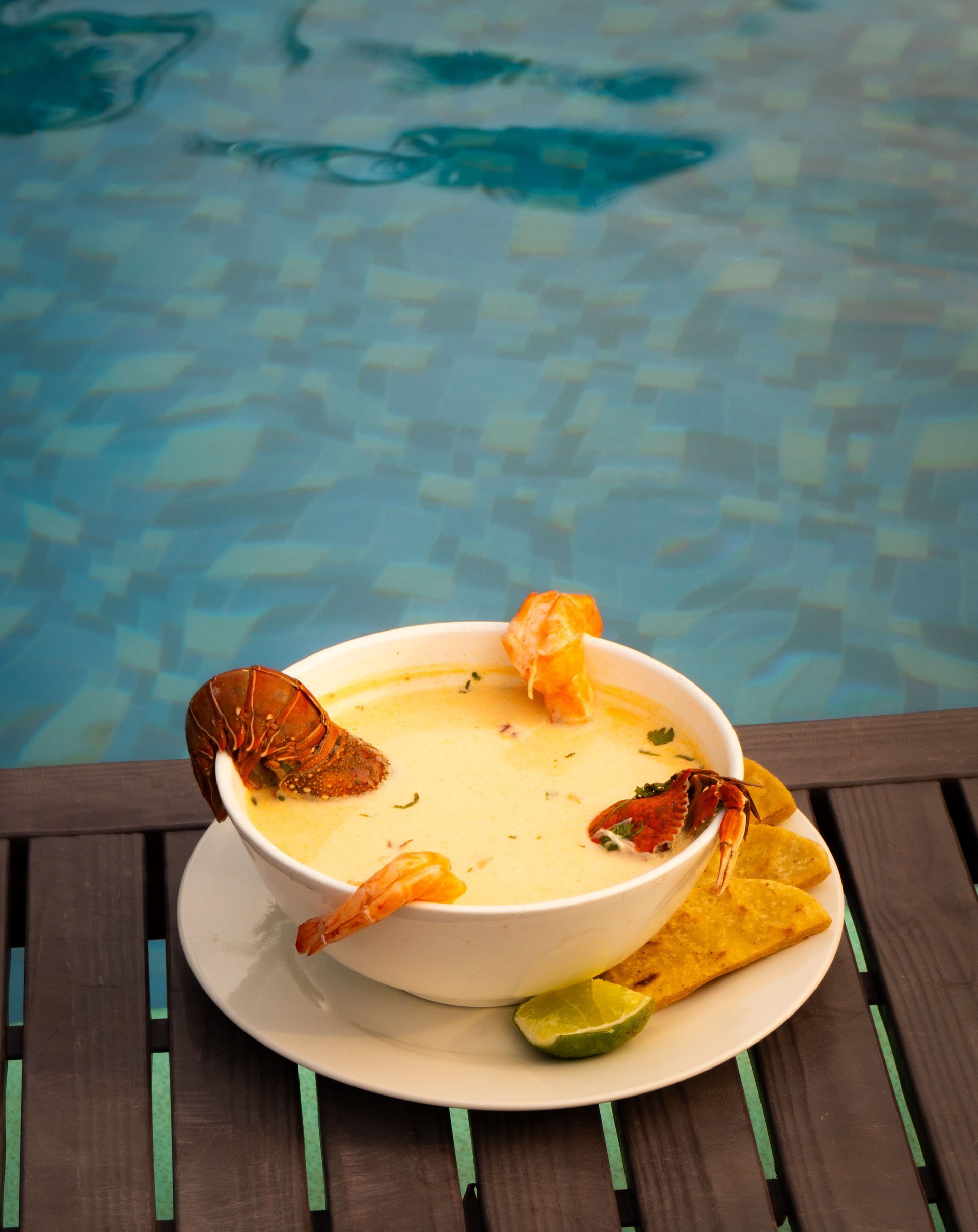 Tazón de sopa cremosa con langosta, camarones y patatas fritas junto a una piscina.