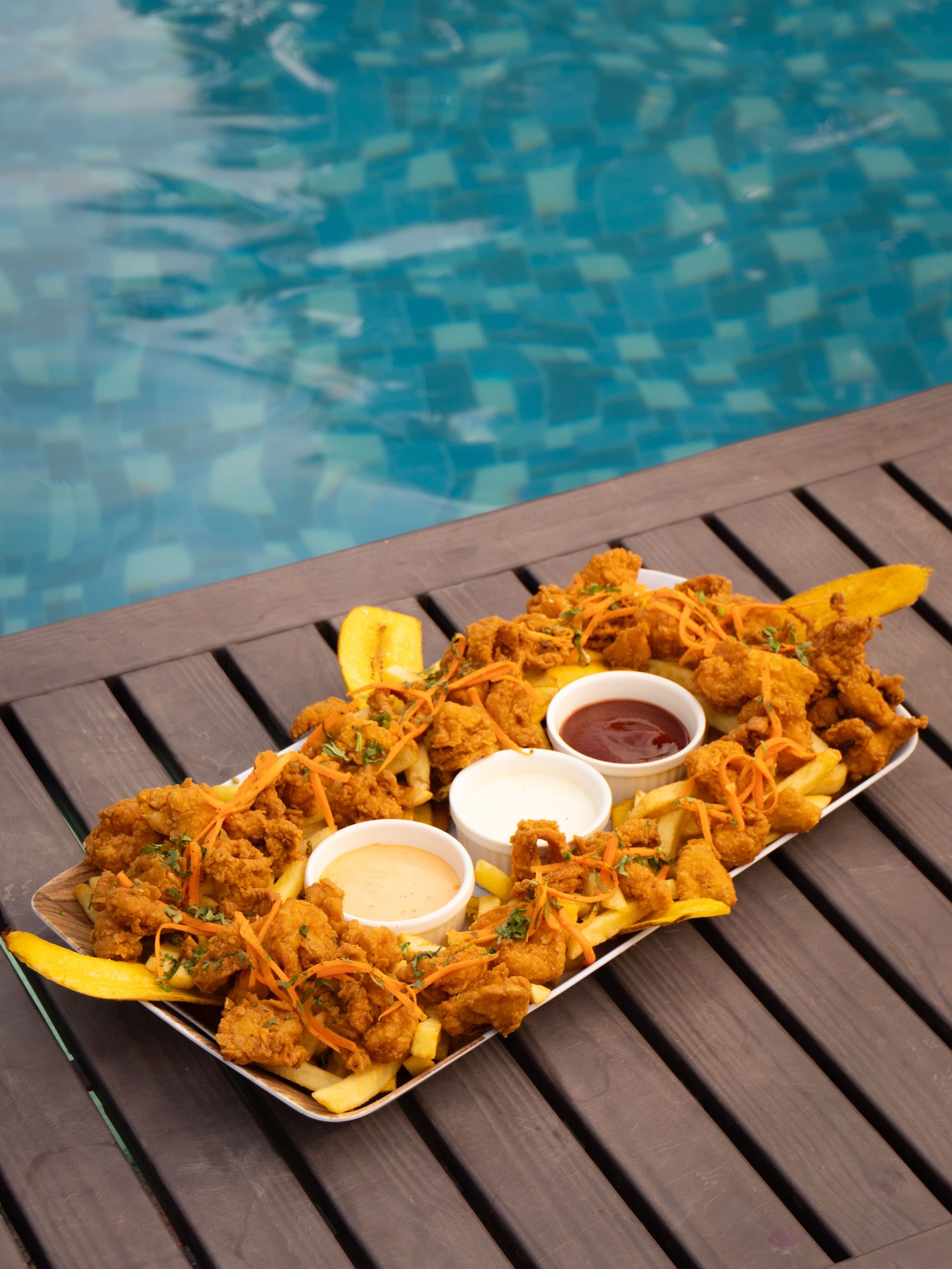 Bandeja de comida frita con tres salsas, cerca de una piscina.
