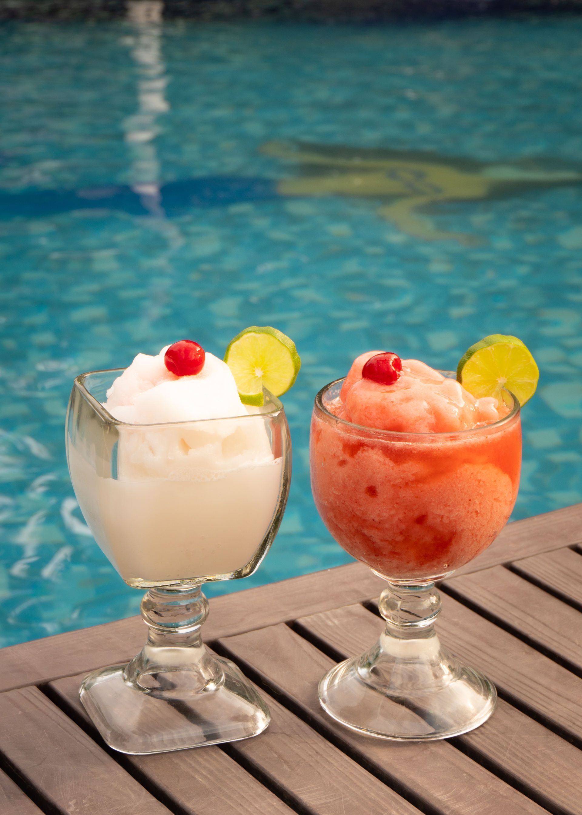 Dos cócteles helados, uno blanco y otro tinto, se sirven junto a una piscina. Cada uno decorado con lima y cereza.