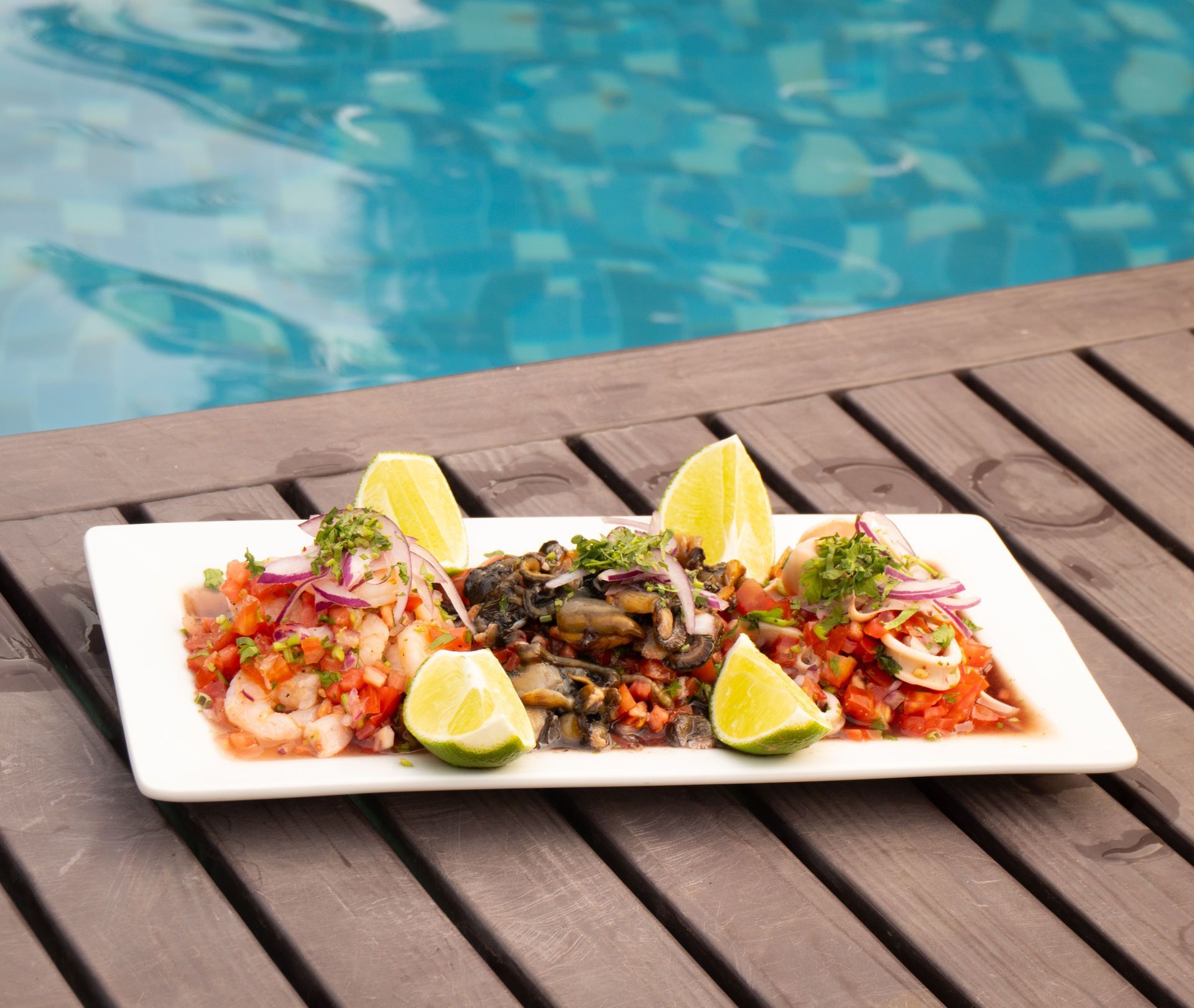 Plato de ceviche de mariscos con gajos de limón junto a una piscina.