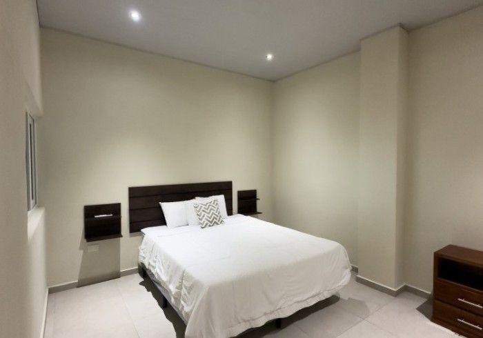 Dormitorio con una cama, dos mesitas de noche flotantes y una cómoda; paredes de color beige claro y ropa de cama blanca.