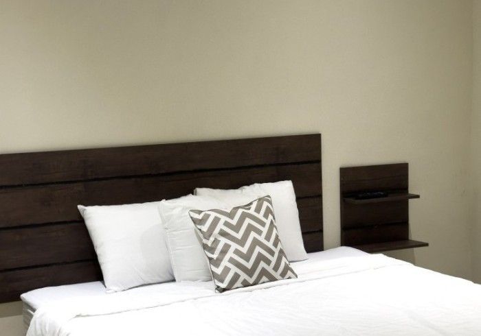 Cama con ropa de cama blanca, cabecero marrón y almohada estampada gris. Pequeña estantería a la derecha.