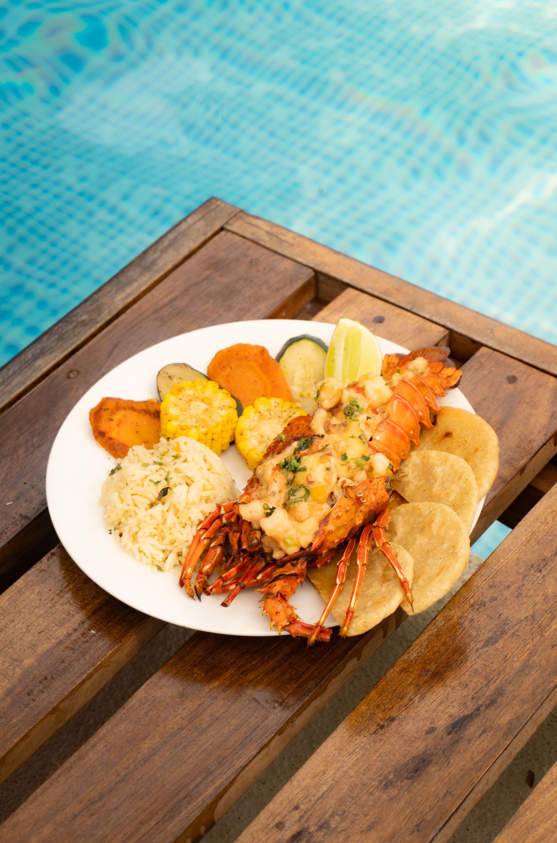 Langosta con guarniciones en un plato colocado sobre una mesa de madera junto a una piscina.