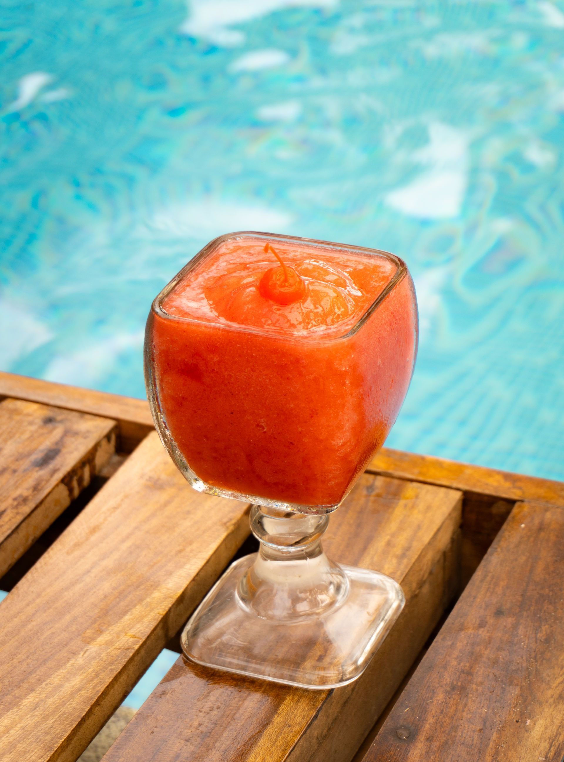 Bebida roja congelada en un vaso cuadrado sobre una terraza de madera, con piscina al fondo.