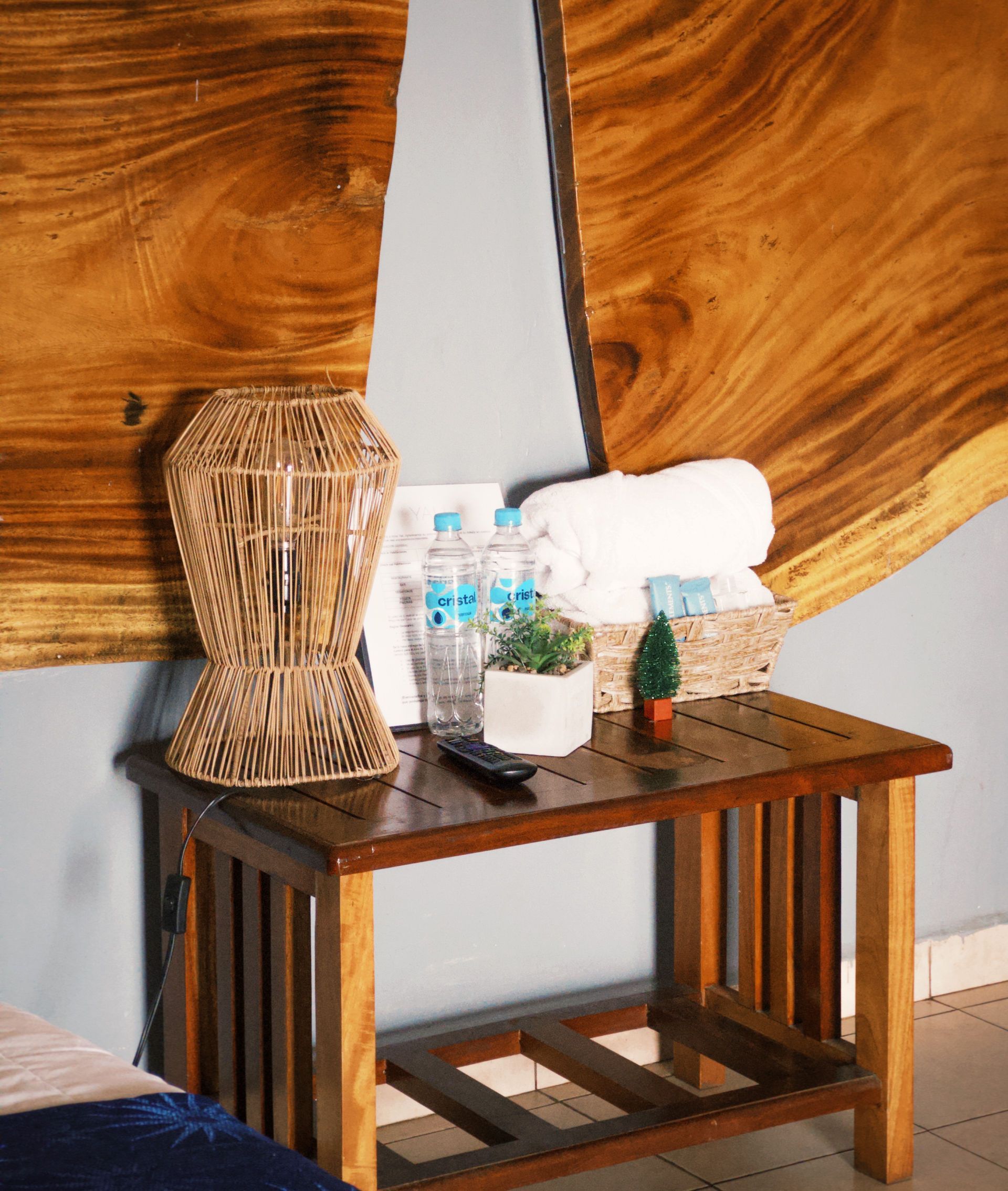 Mesita de noche de madera con lámpara, botellas de agua, toallas y una pequeña planta contra una pared revestida de madera.