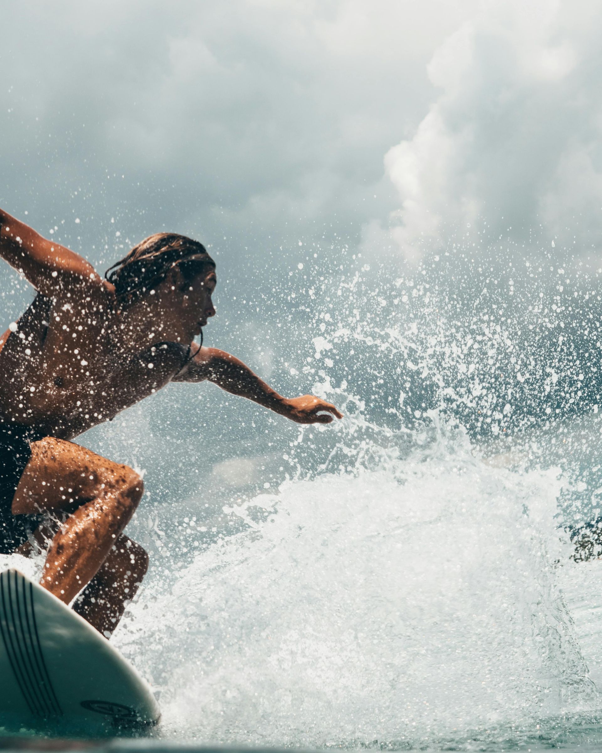 Surfista montando una ola, con el brazo extendido, salpicando agua, contra un cielo nublado.