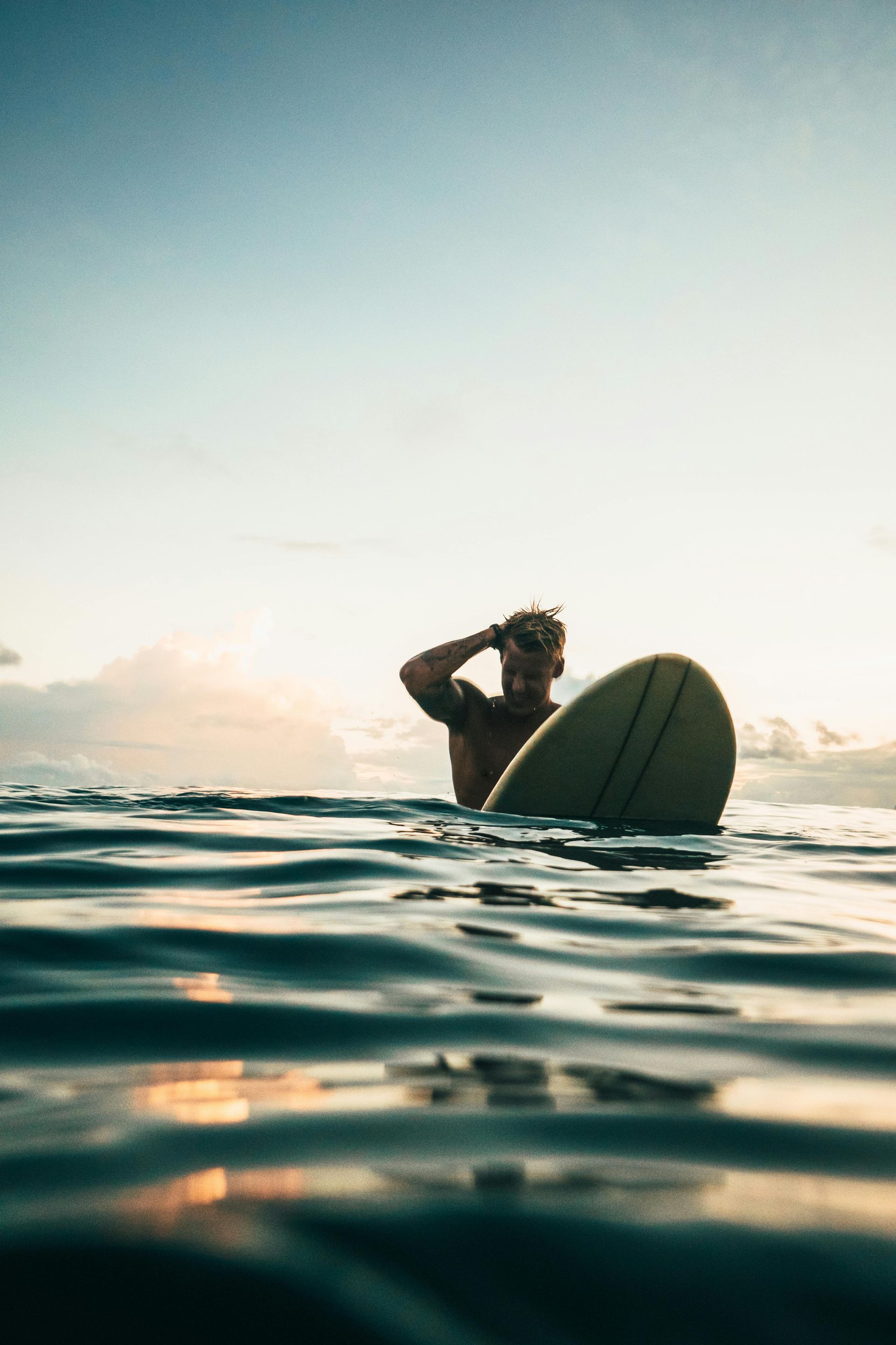 Hombre en el océano, sosteniendo una tabla de surf, mirando hacia el cielo al atardecer.