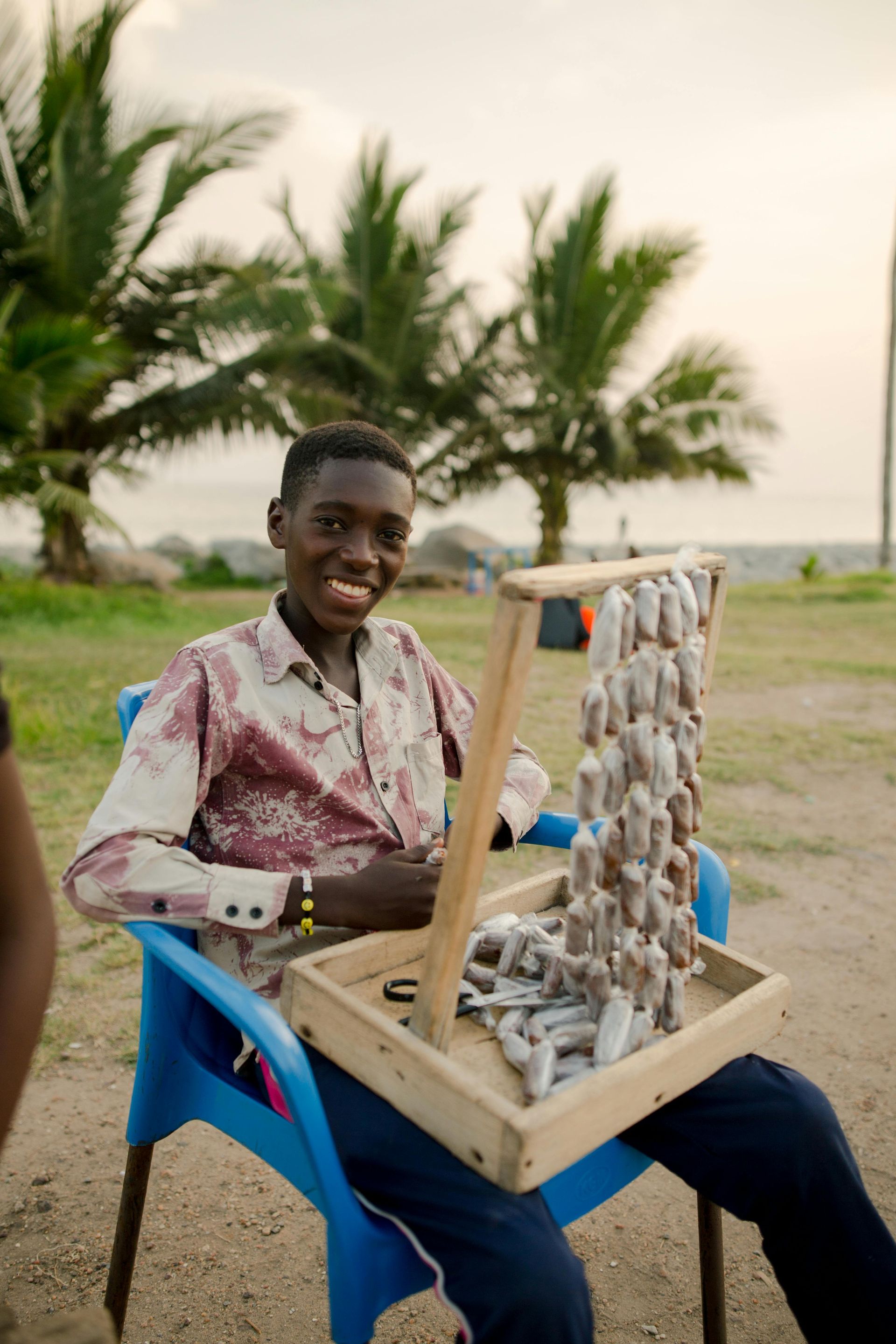 Joven sonriente vendiendo joyas al aire libre, sentado en una silla azul; palmeras al fondo.