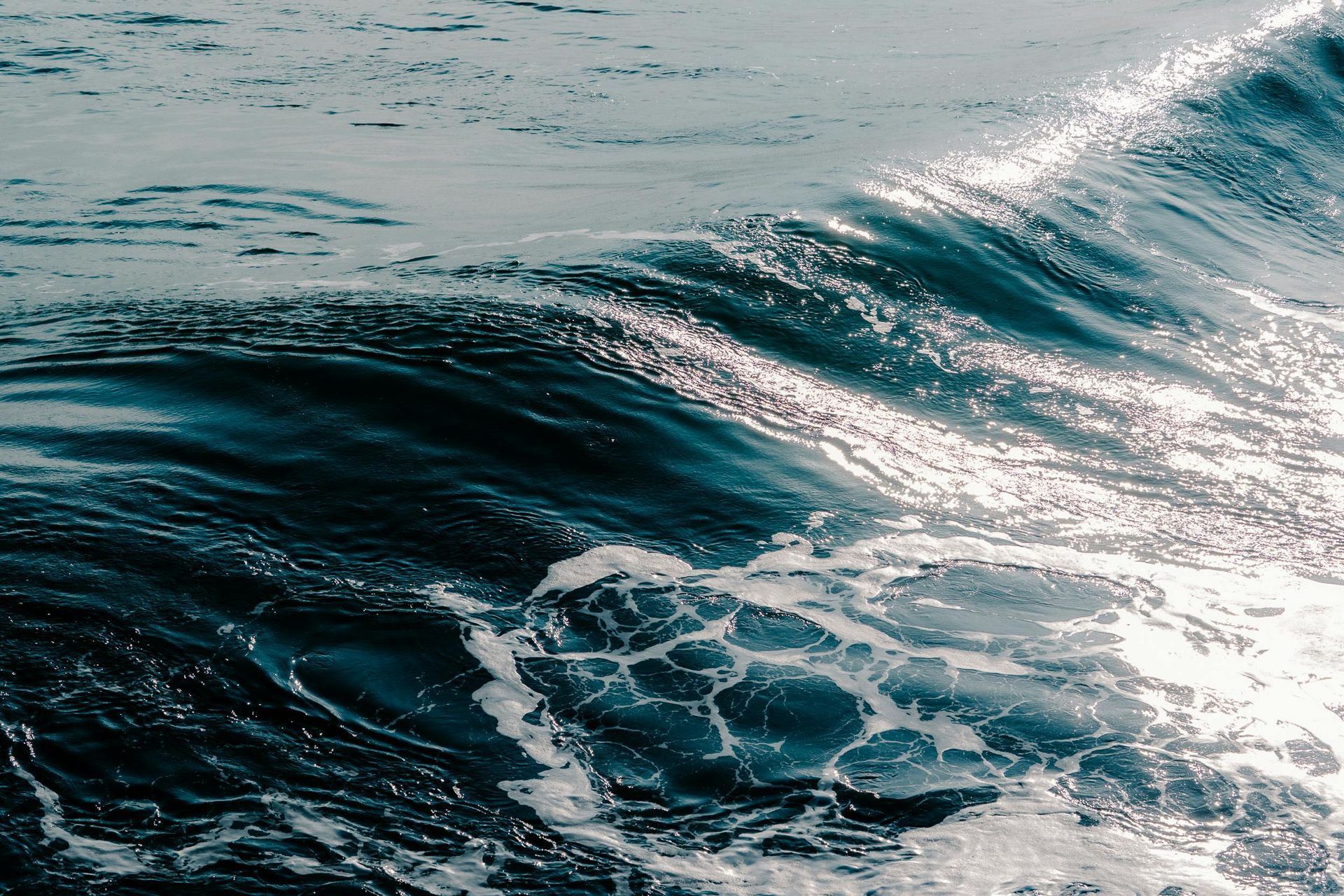 Olas del océano rompiendo, superficie iluminada por el sol. Agua azul oscuro con espuma blanca.