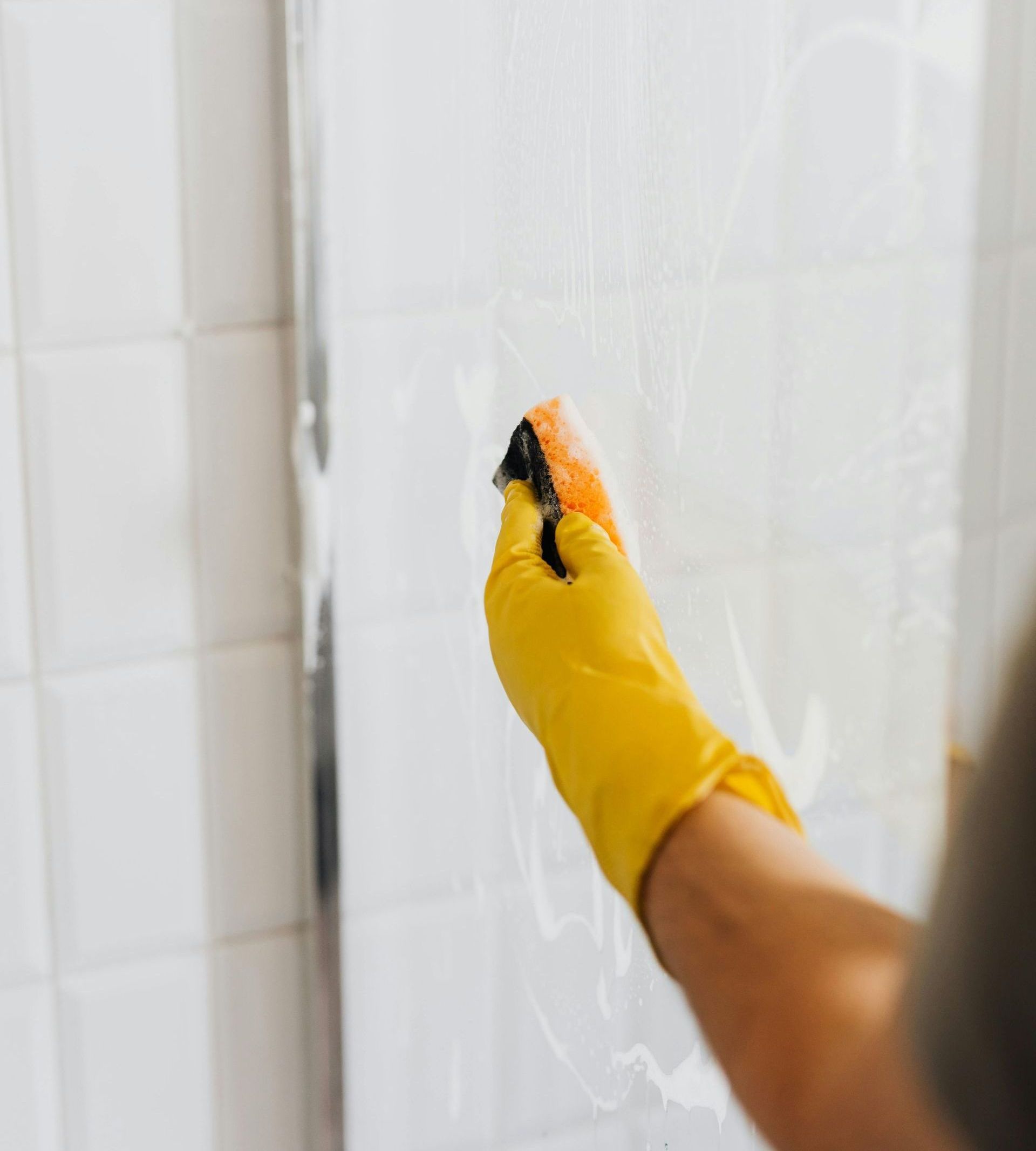 A person wearing yellow gloves is cleaning a shower door with a sponge.