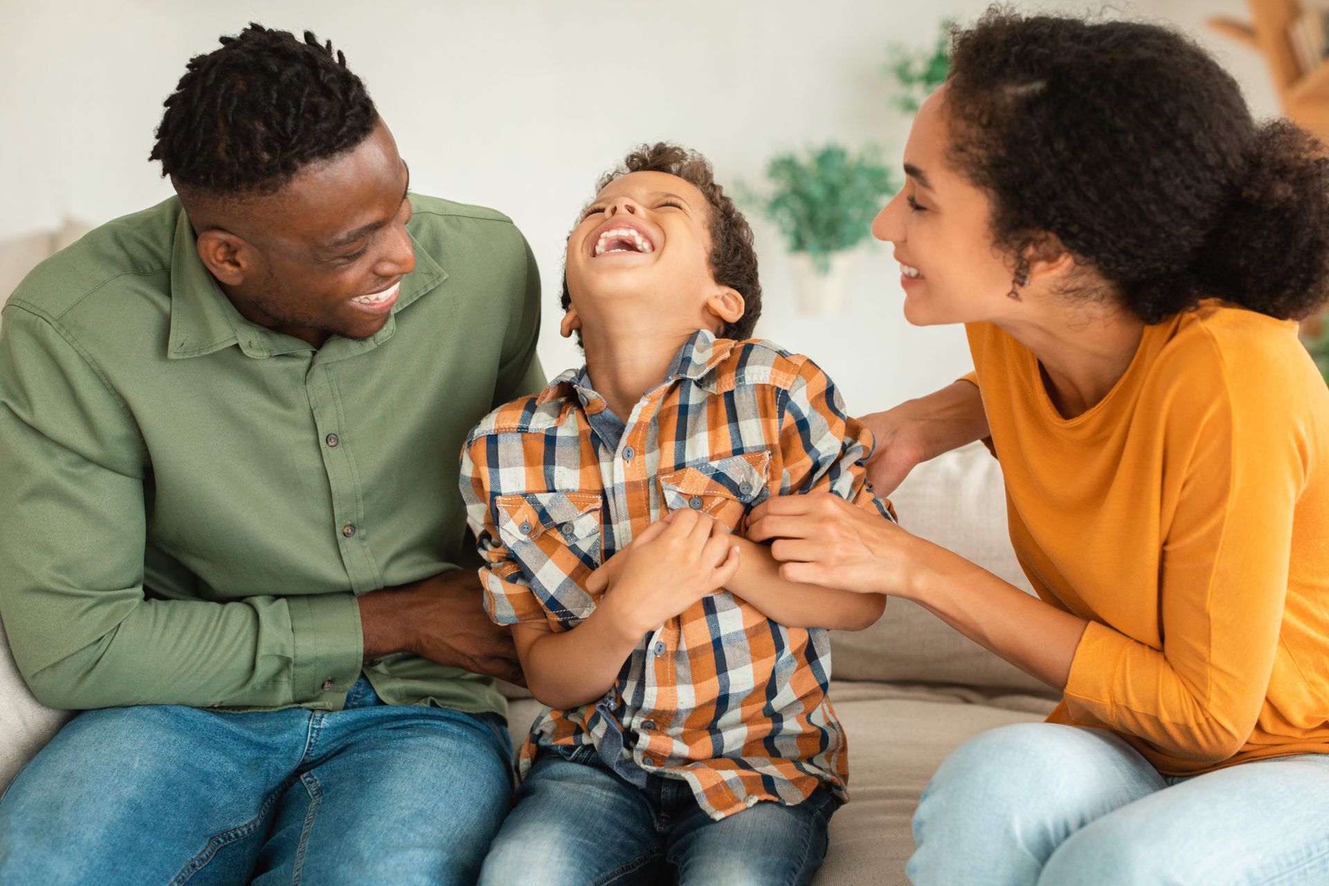 Parents sit on a sofa, tickling their laughing child between them.