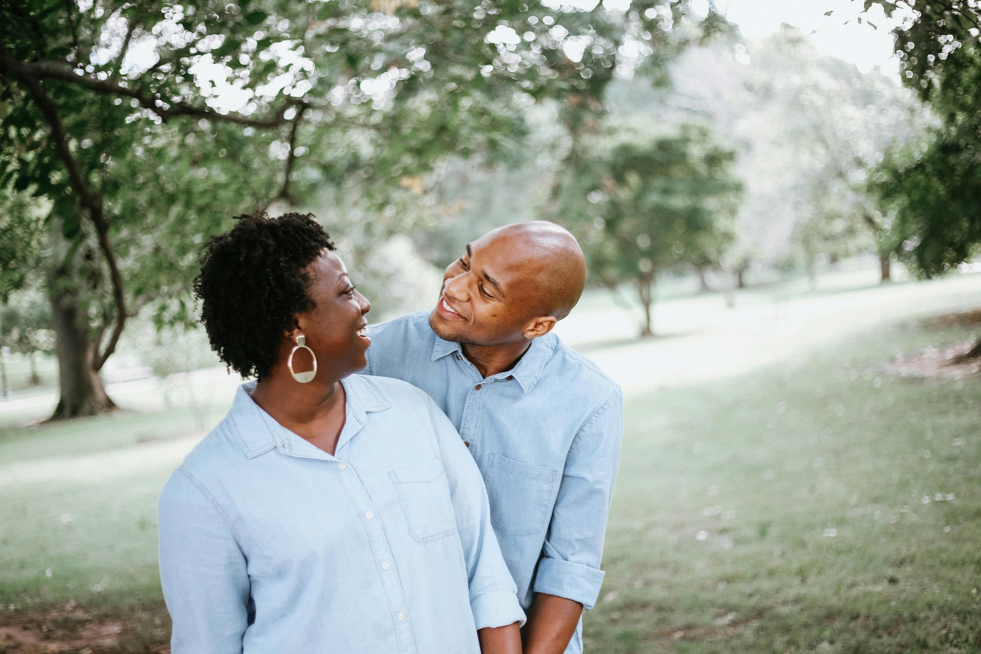 Couple smiles at each other in a park, blue shirts, trees in the background.