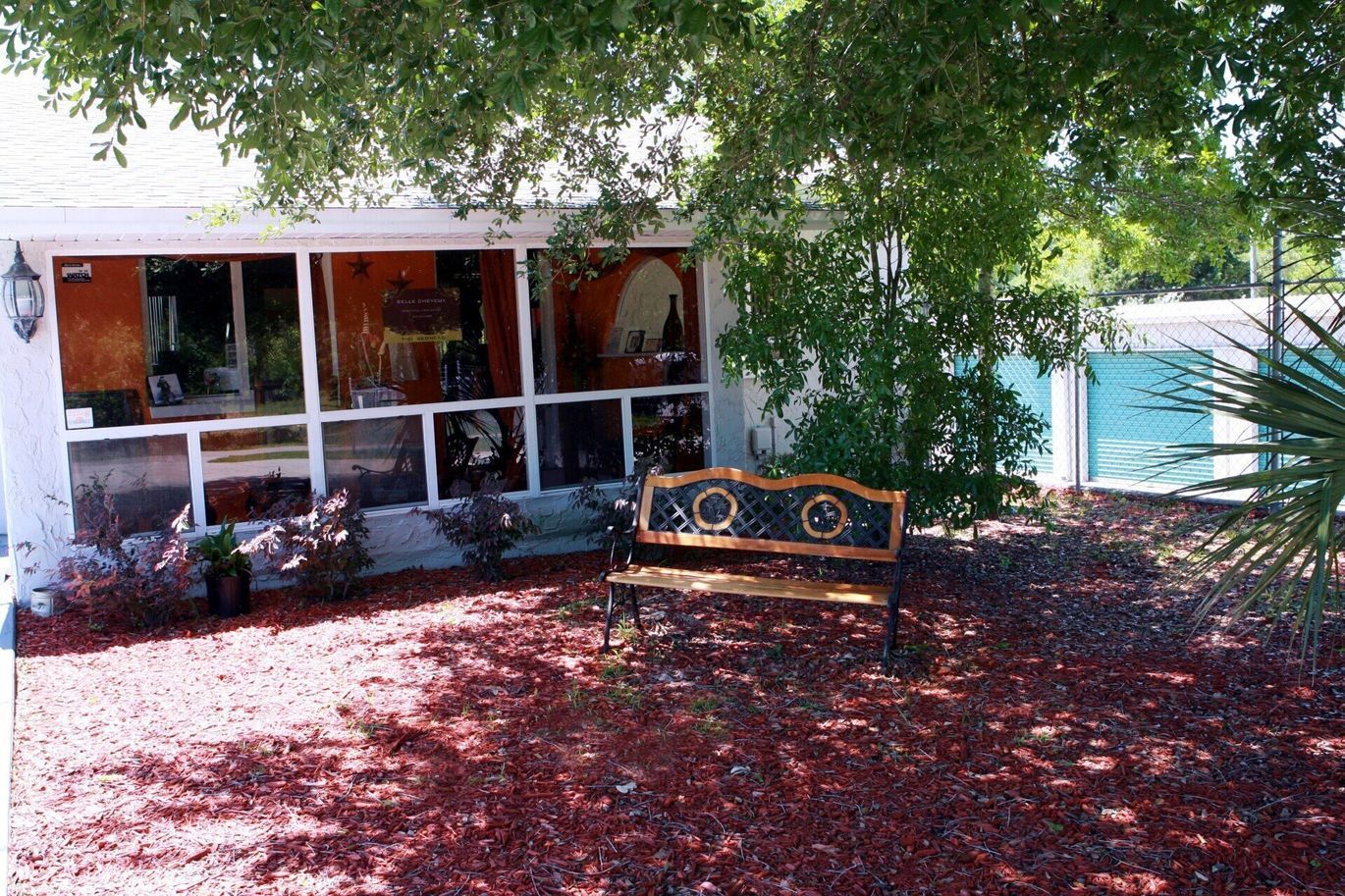 Bench in front of a porch with screened windows, covered in red mulch and shaded by a tree.