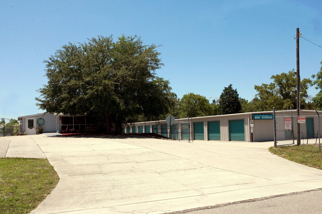 Storage units with a paved lot and a tree under a clear, blue sky.