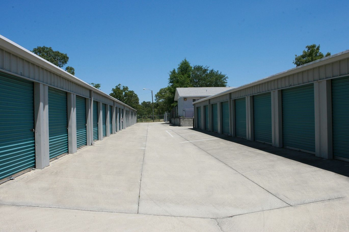 Rows of teal storage unit doors face each other; a concrete walkway divides them under a clear, blue sky.
