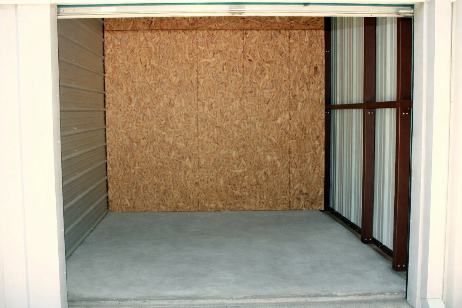Empty storage unit with concrete floor, metal and wood walls, and open door.