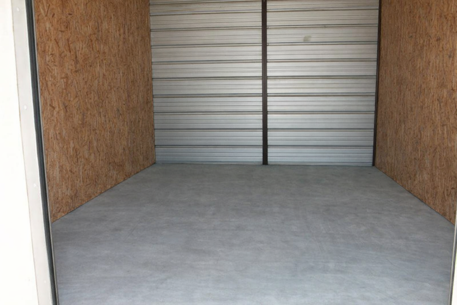 Empty storage unit with gray concrete floor, corrugated metal door, and wood-paneled walls.