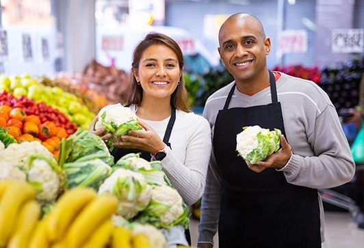 Two smiling grocery store employees holding cauliflower. Produce shelves in the background.