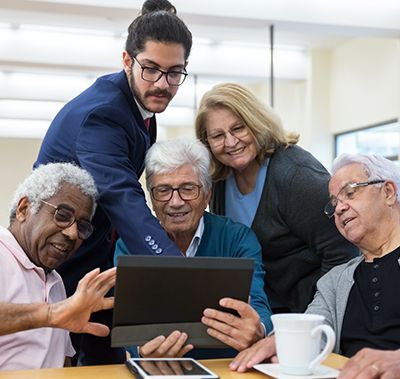 Young man helps group of seniors view tablet screen, smiling in a well-lit room.