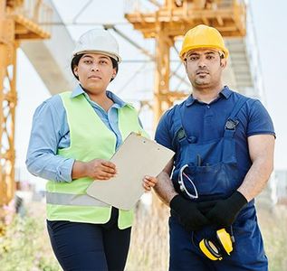 Construction workers in hard hats: woman with clipboard, man with earmuffs, on site.