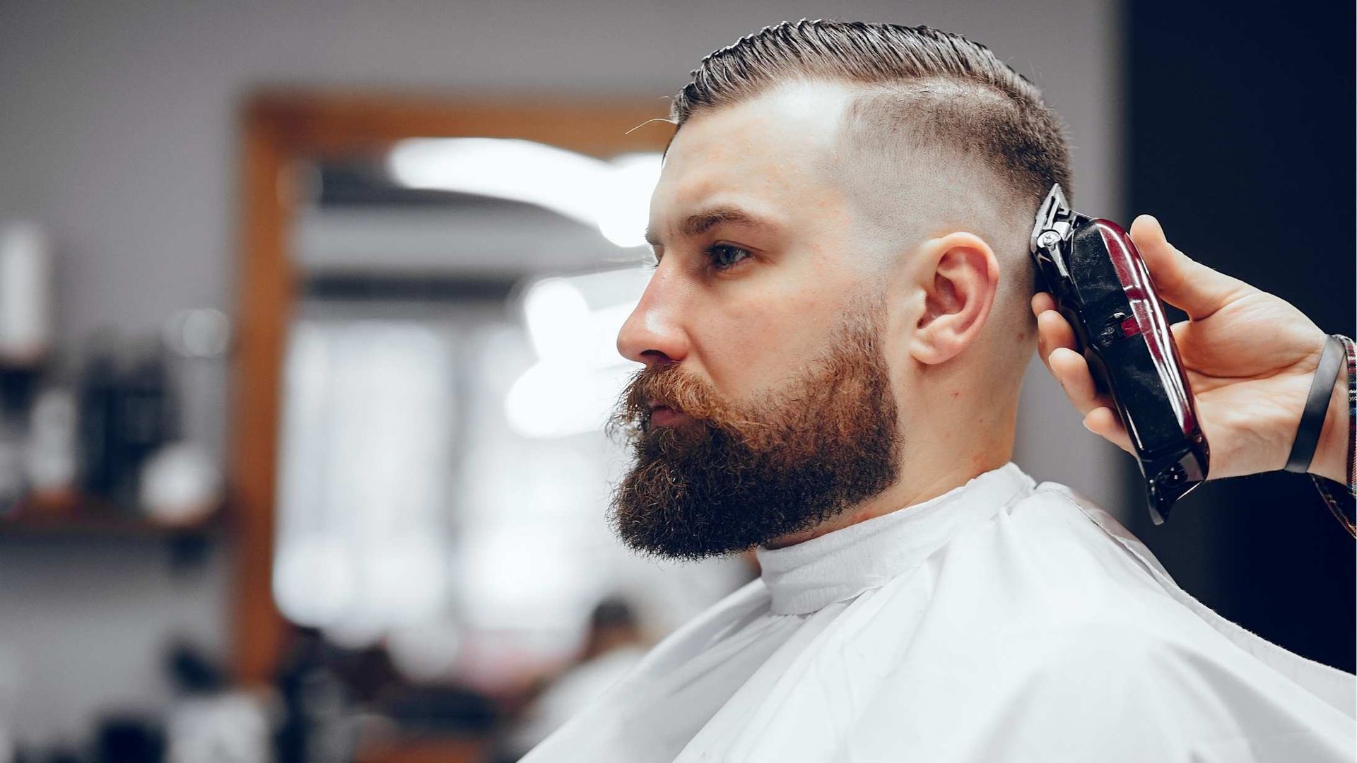 Barber trimming a client's hair with clippers in a salon. The client has a full beard and is wearing a white cape.