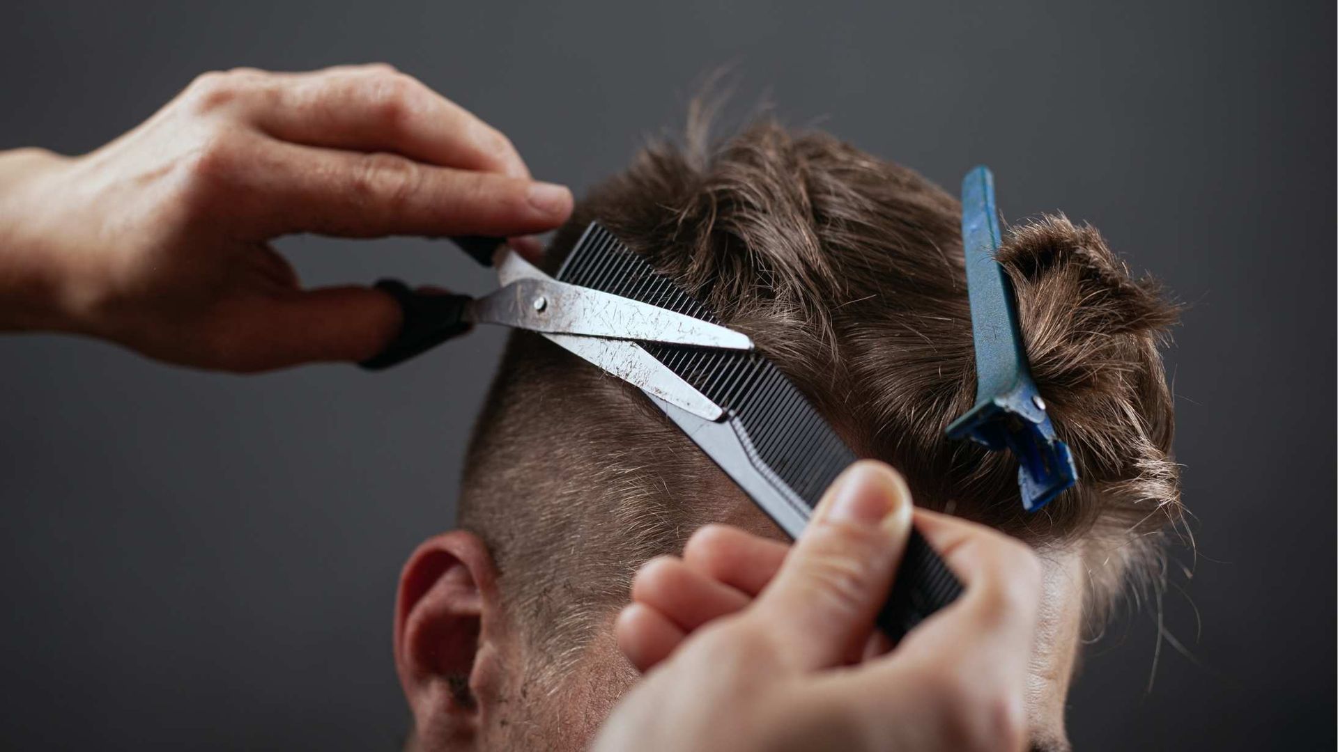 Hairdresser trimming a person's hair with scissors and comb against a gray background.