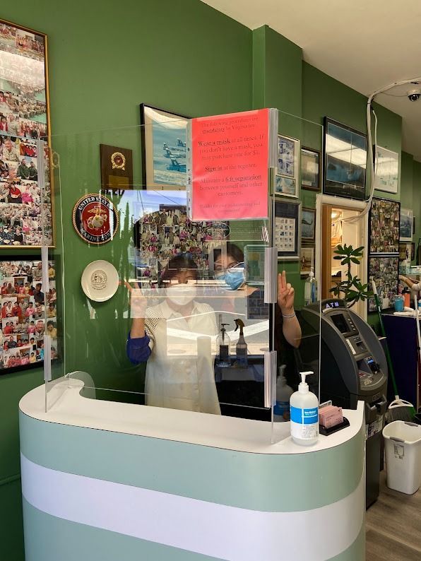 Two masked people behind a protective barrier at a salon's front desk. One person gives a thumbs up.
