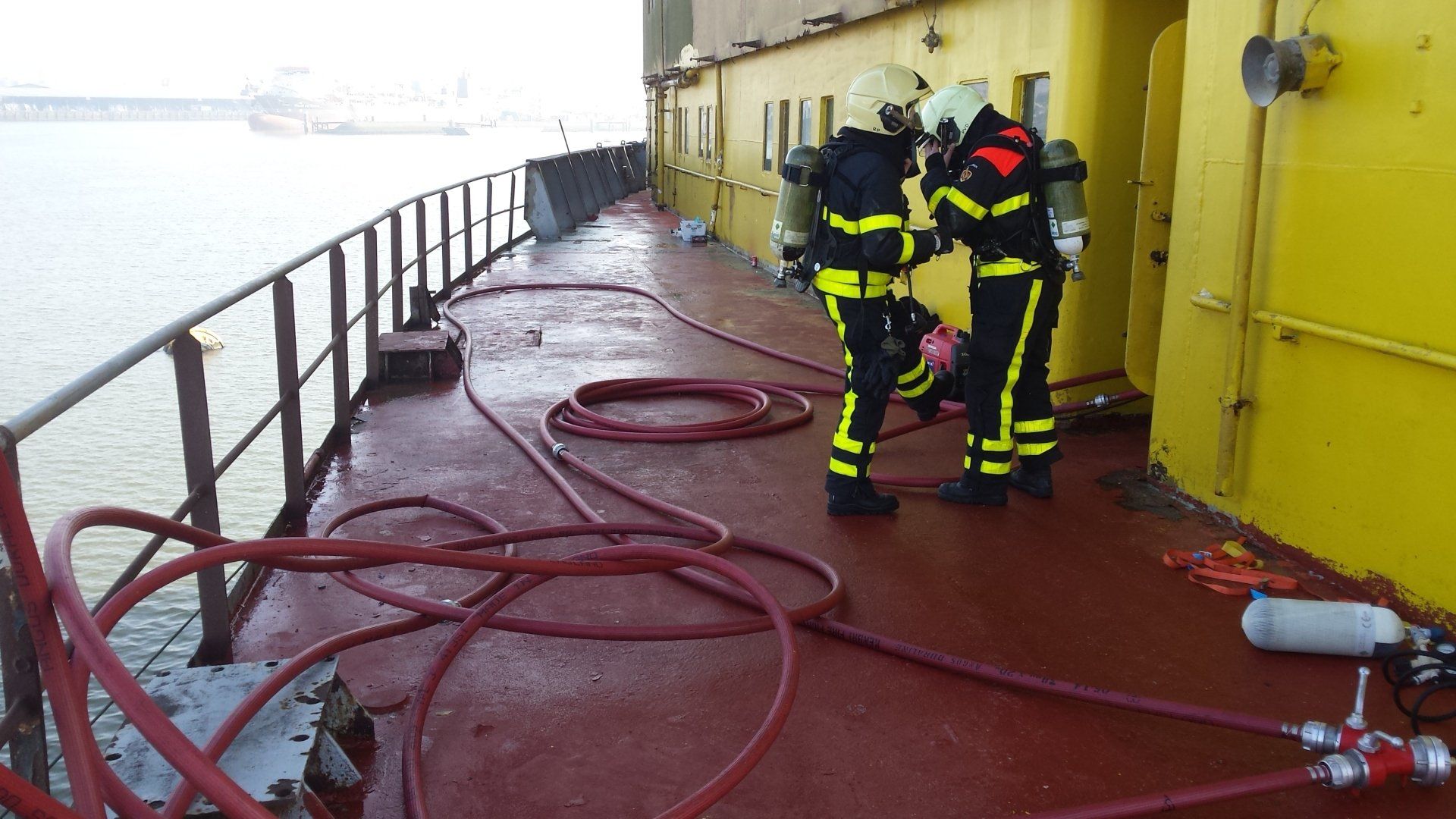 Brandweerlieden in gele kleding op het dek van een schip, die bezig zijn met slangen.