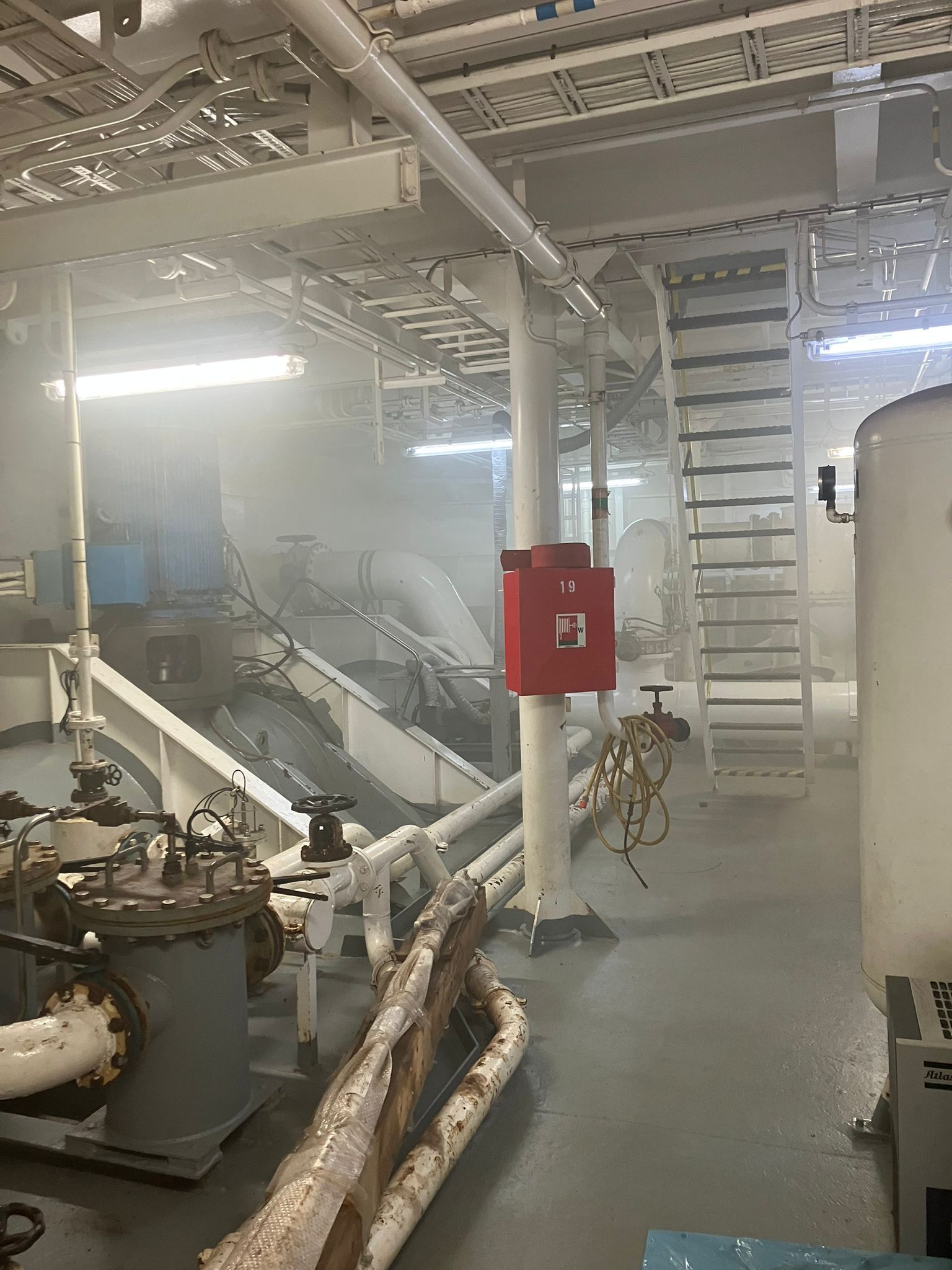 Interieur van de machinekamer van een schip, gevuld met mist. Rode brandslangenkast op een witte zuil; ladders en machines zichtbaar.