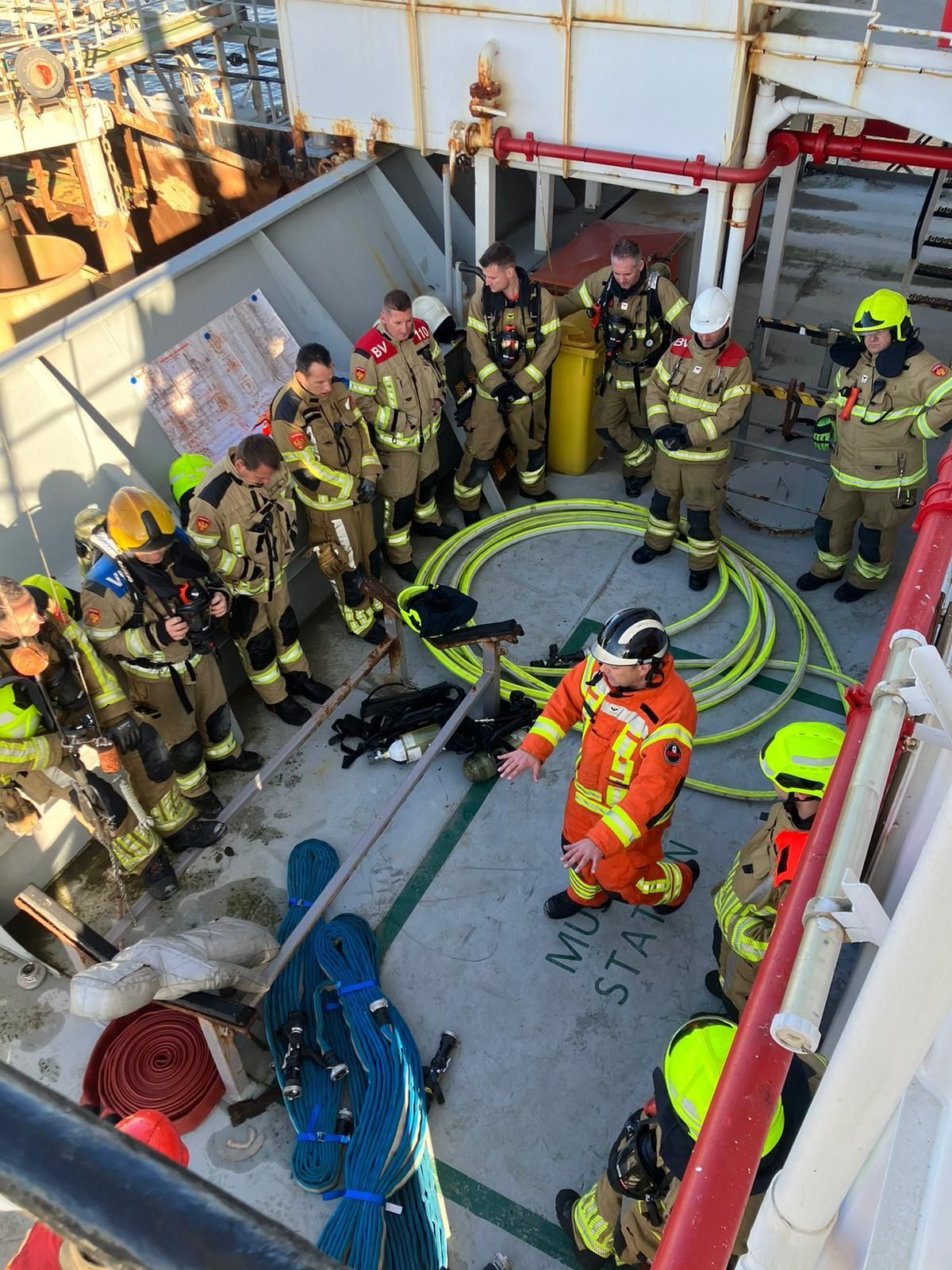 Brandweerlieden in uniform op een dek luisteren naar een instructeur. Gele slangen, rode leidingen en blauwe touwen zijn zichtbaar.