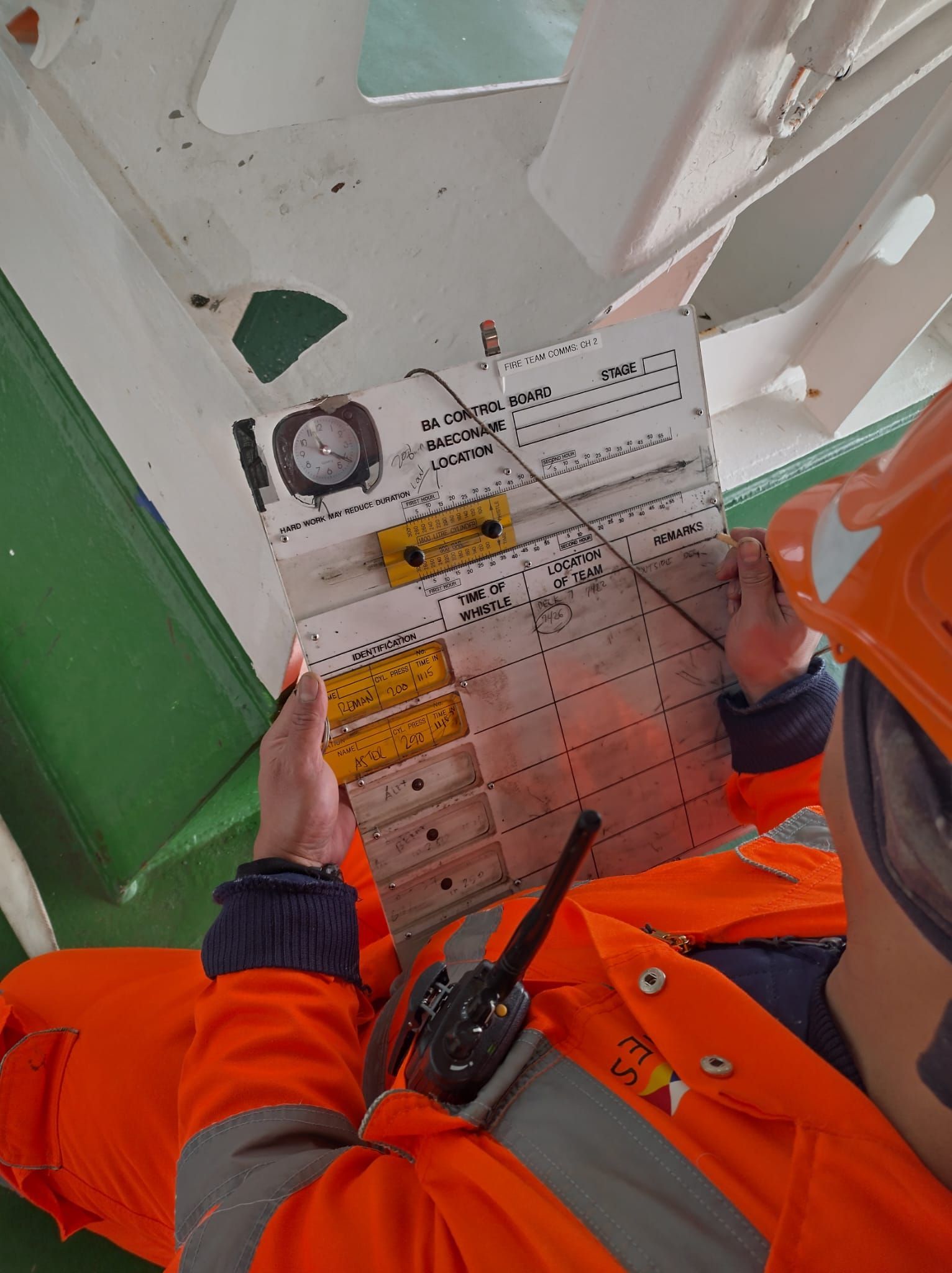 Persoon in oranje veiligheidsuitrusting inspecteert een witte kaart die aan een balk is bevestigd. Een kompas en radio zijn zichtbaar.