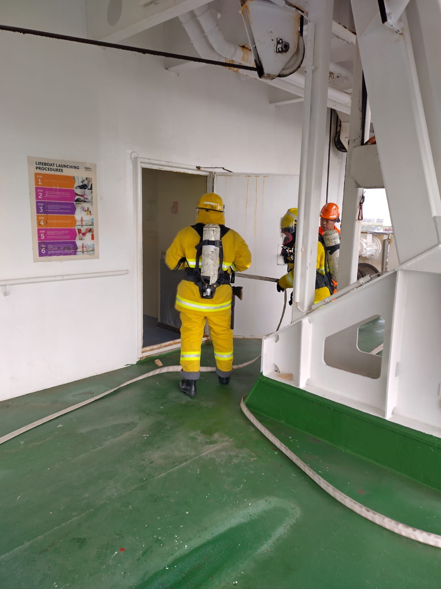 Brandweerlieden in gele pakken en met ademhalingsmaskers gaan een deuropening van een schip binnen, met een slang in hun hand.
