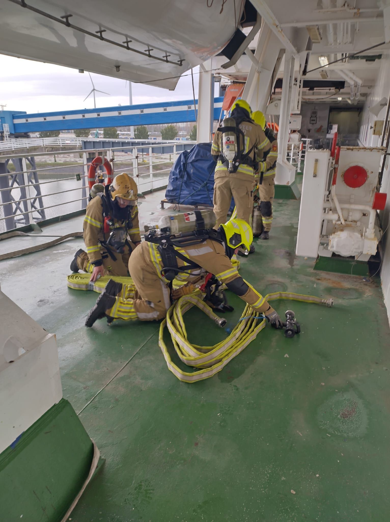 Brandweerlieden in beschermende kleding op het dek van een schip, bezig met een slang.