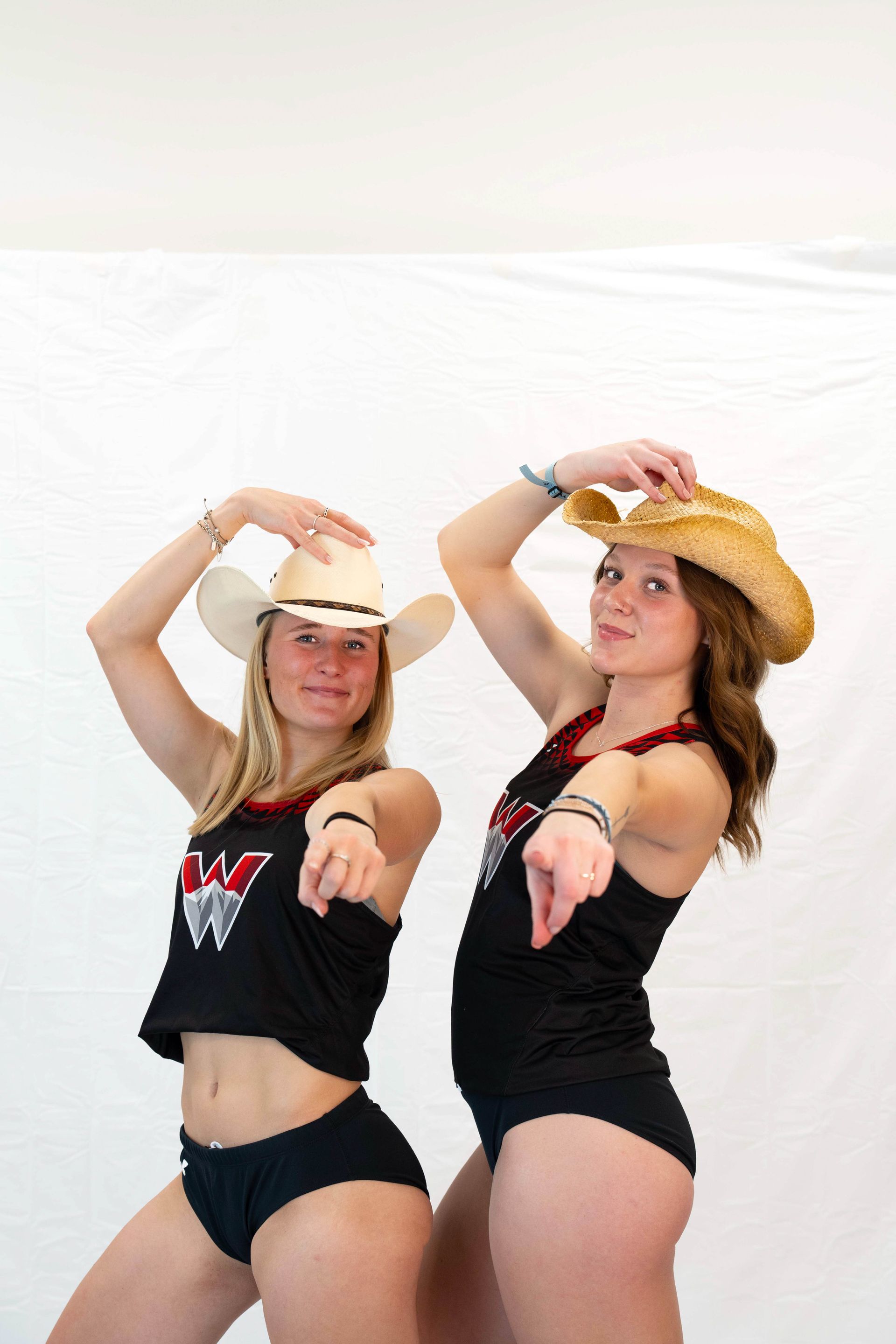Two women in cowboy hats and black tank tops point forward.