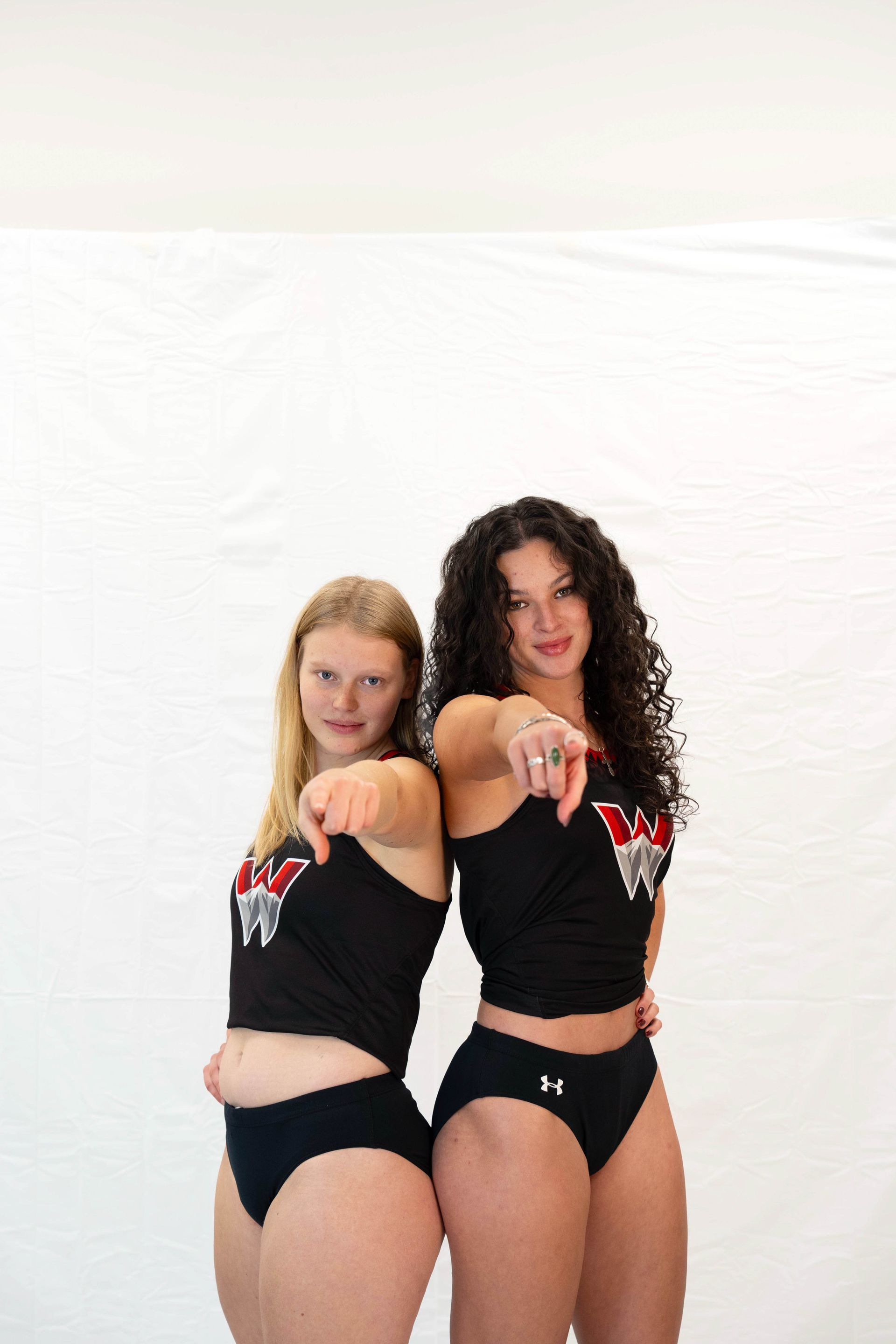 Two women in black athletic wear point forward against a white backdrop.