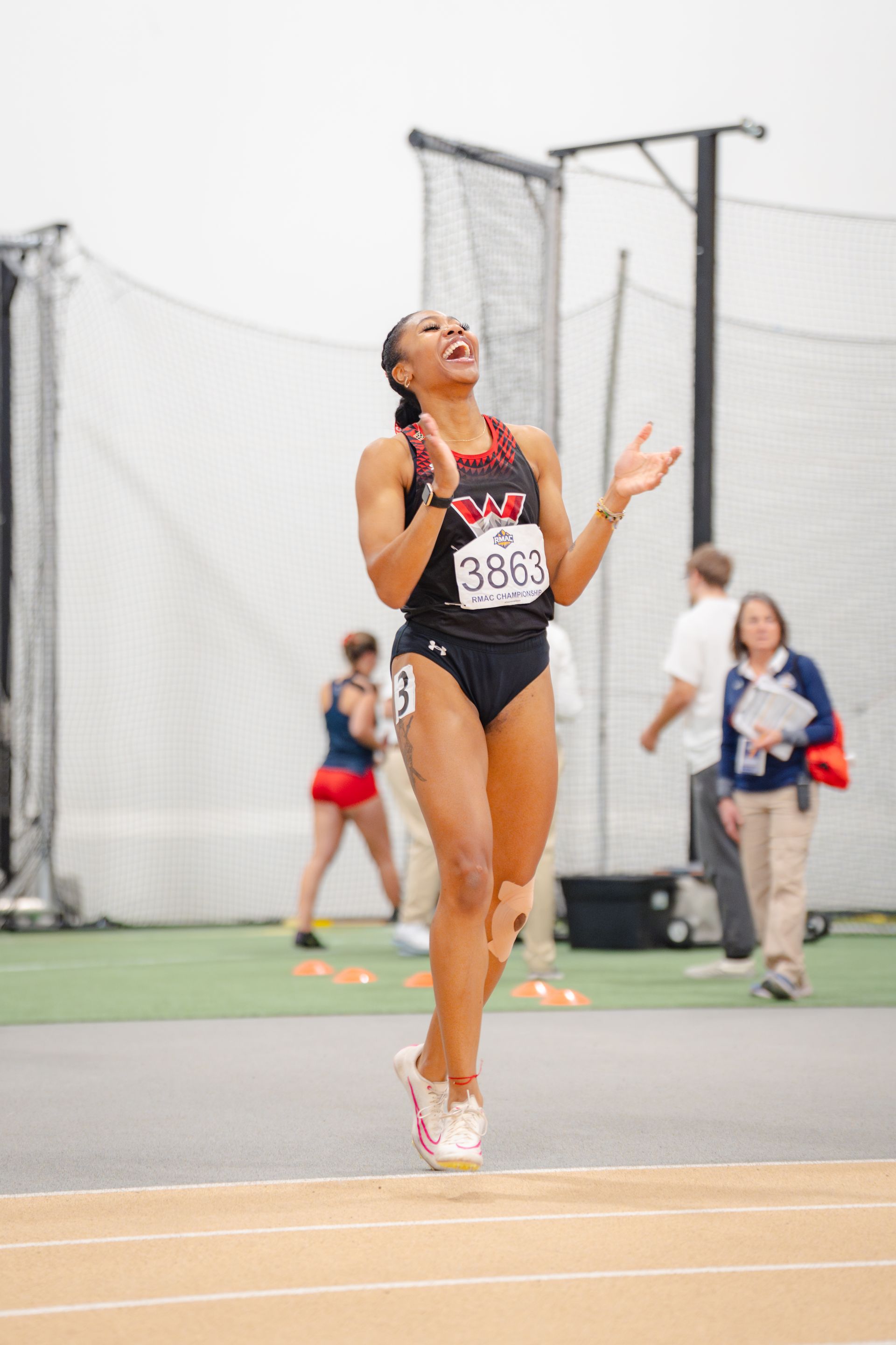 A female track athlete in a black uniform celebrates a victory, clapping and smiling, indoors on a track.