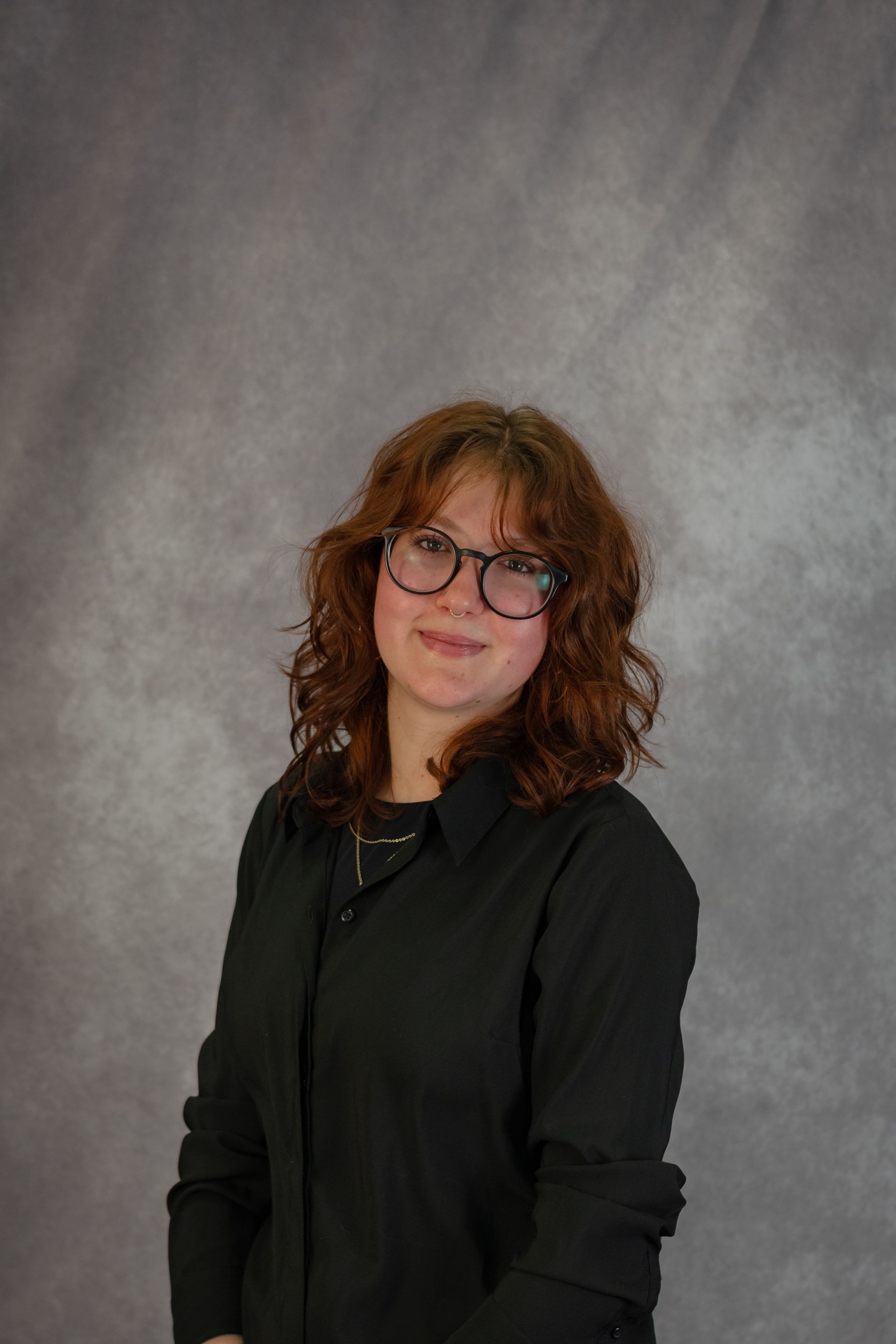 Woman with red hair wearing glasses and a black shirt, smiling, in front of a mottled gray backdrop.