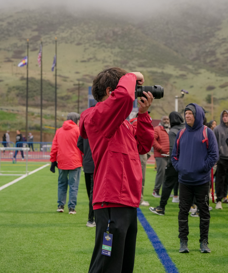 Kai Vong in red jacket taking photos on a sports field with people in the background; mountains in mist.