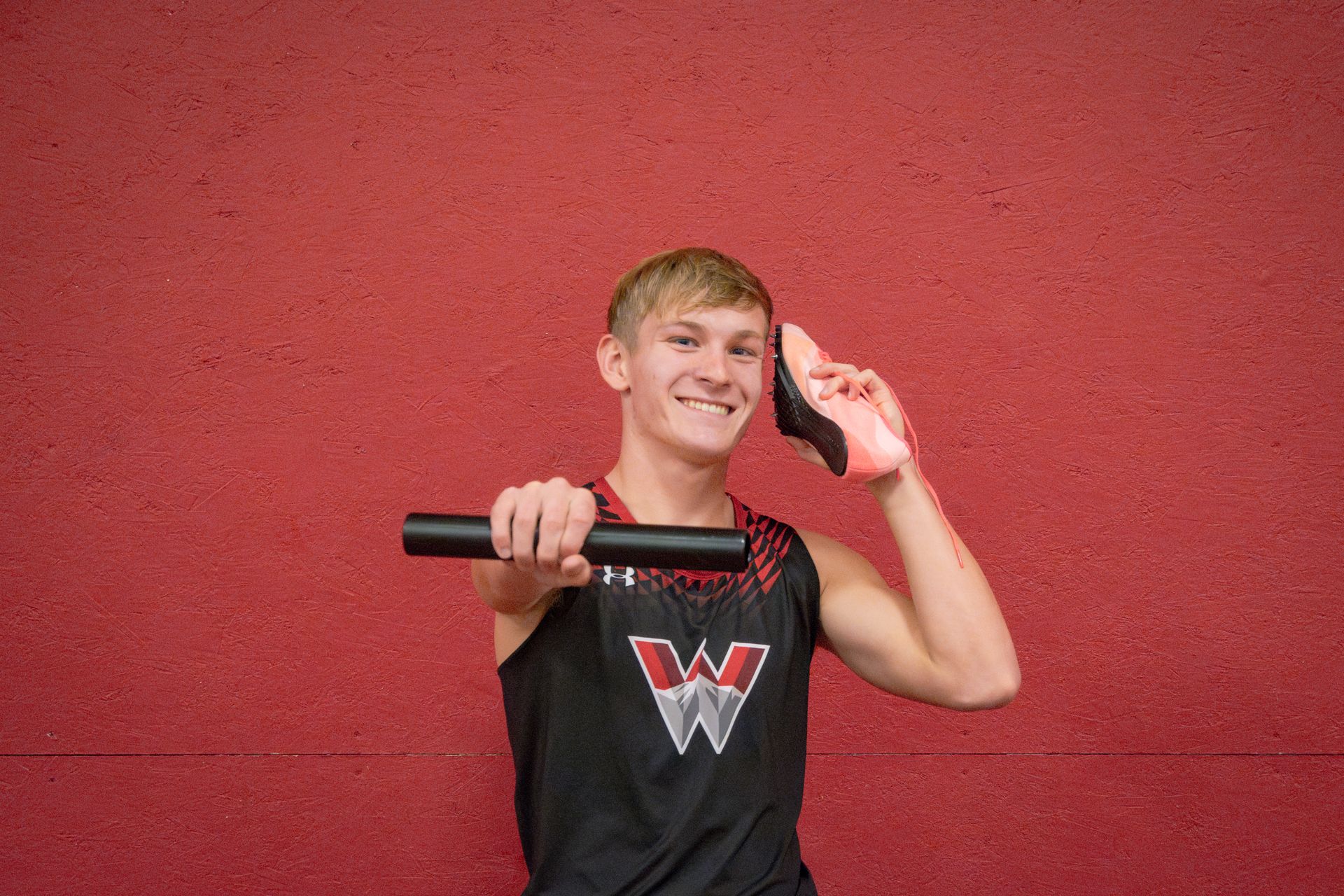 Young track athlete holds a baton and a shoe. Red wall background. He smiles.
