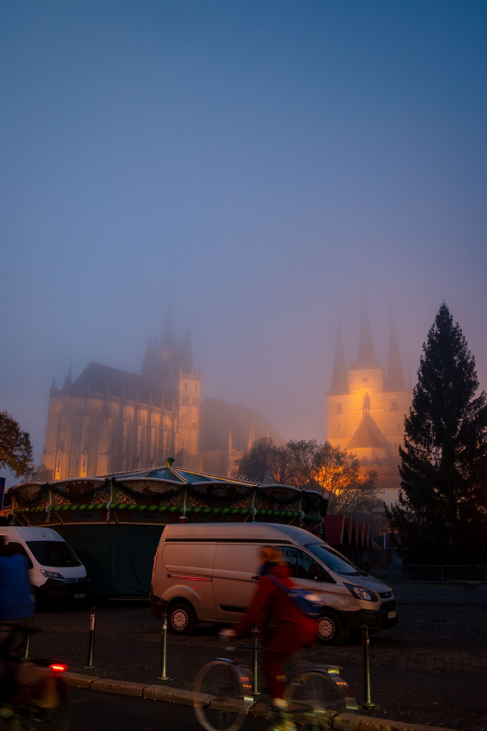 Foggy city scene with obscured cathedral spires, cyclist, and parked vehicles.