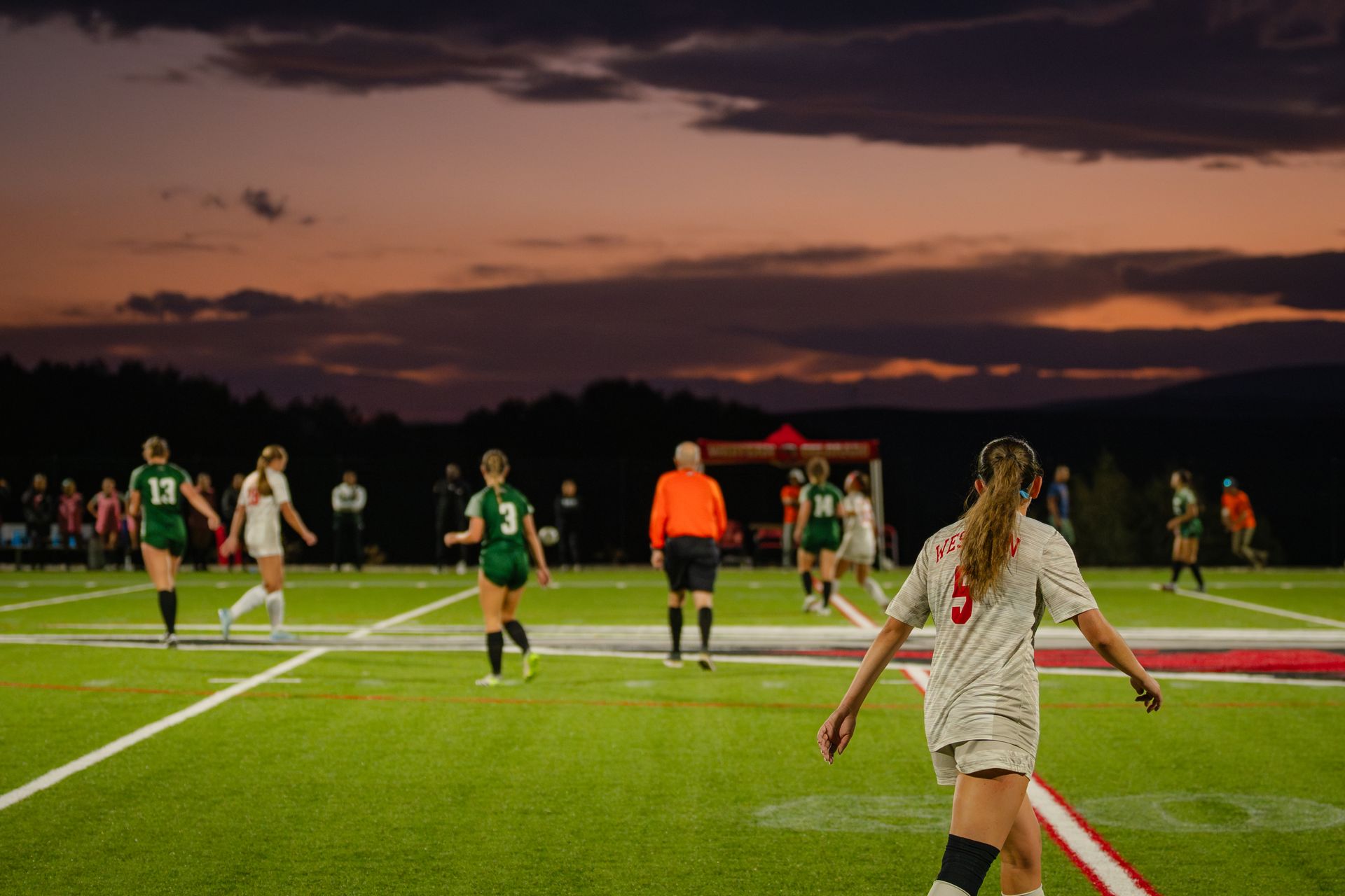Women's soccer game at dusk. Players in green and white uniforms on a field with referee. Sunset in the background.