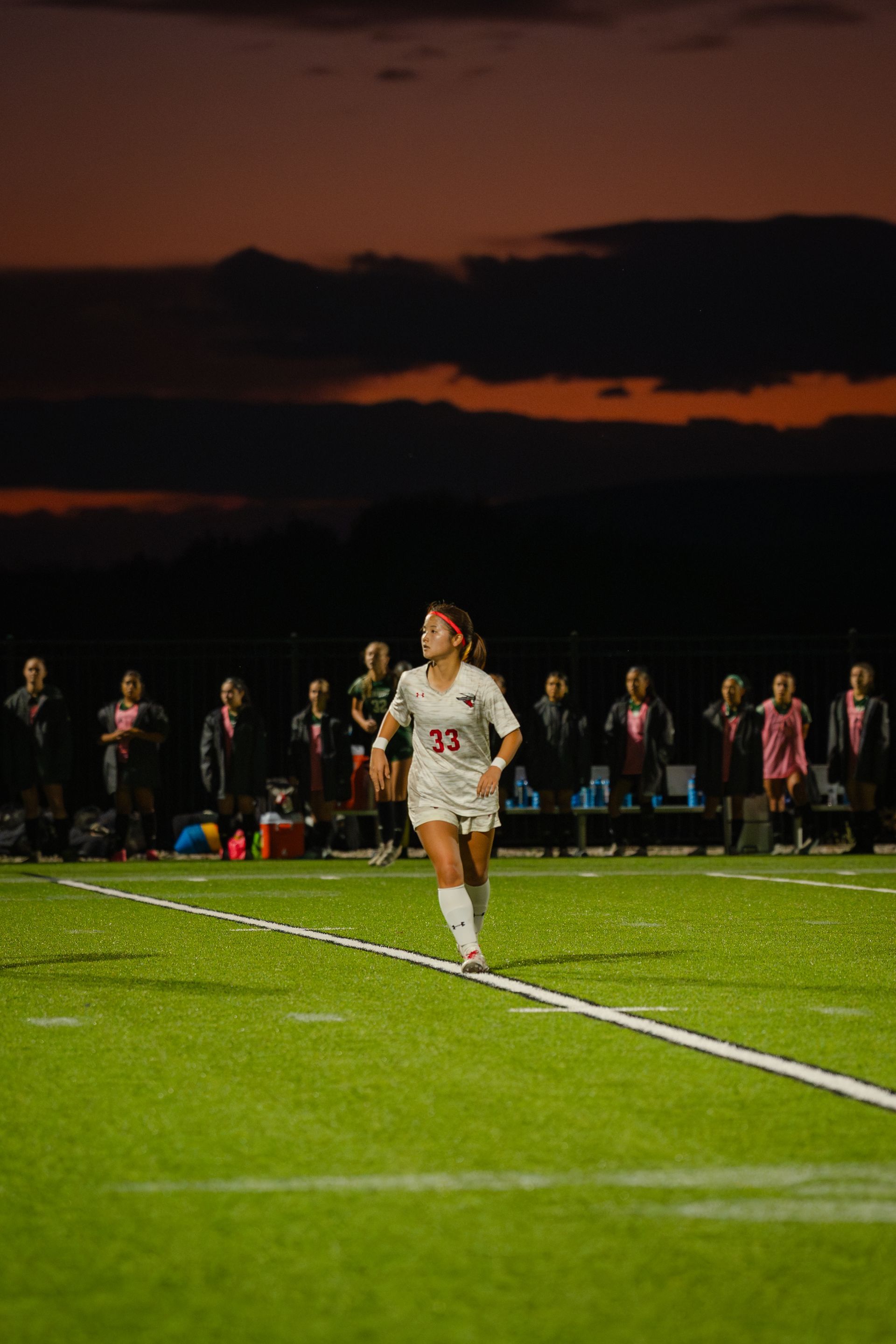 Soccer player in white uniform runs on green field, crowd in background, dusk sky.